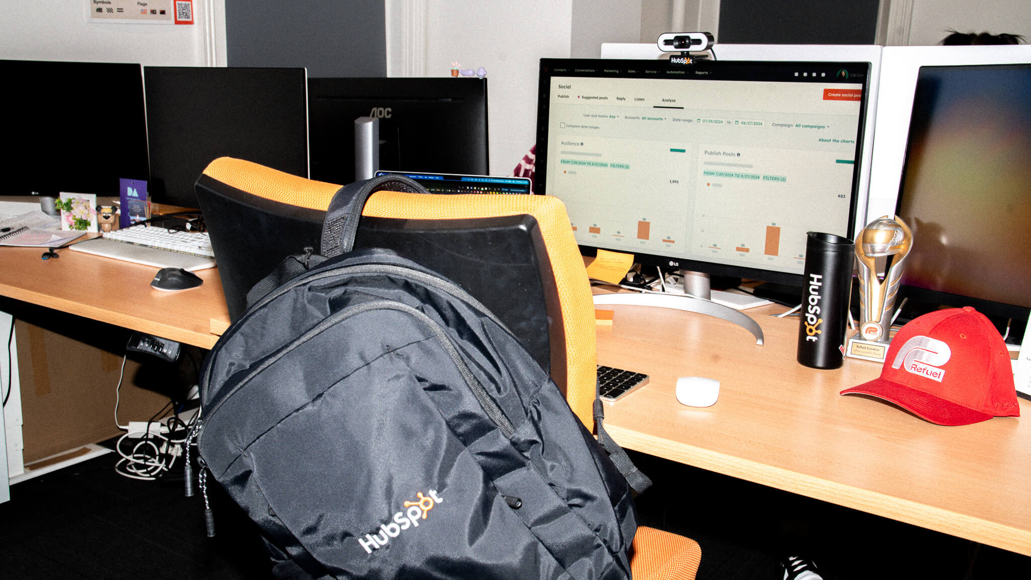 An office desk with a HubSpot backpack on the chair, a HubSpot tumbler, a Refuel cap, and a trophy next to a computer displaying HubSpot analytics.