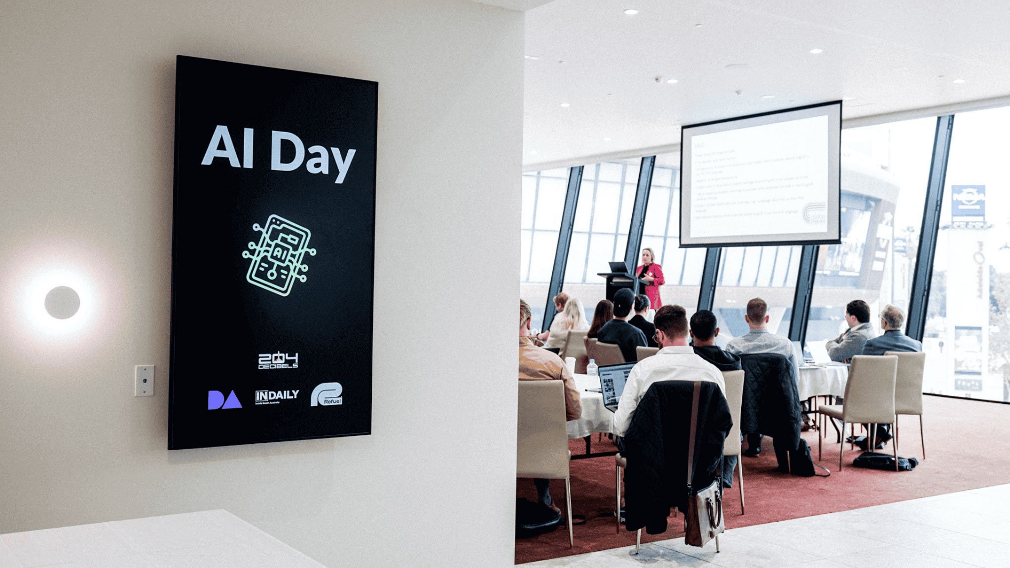 "AI Day" conference sign on wall beside meeting room with attendees watching presenter at projection screen.