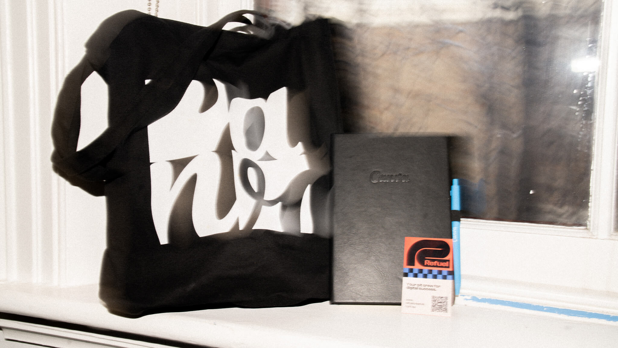 A motion-blurred photo of Canva and Refuel branded merchandise, including a tote bag and notebook, sitting on a windowsill.