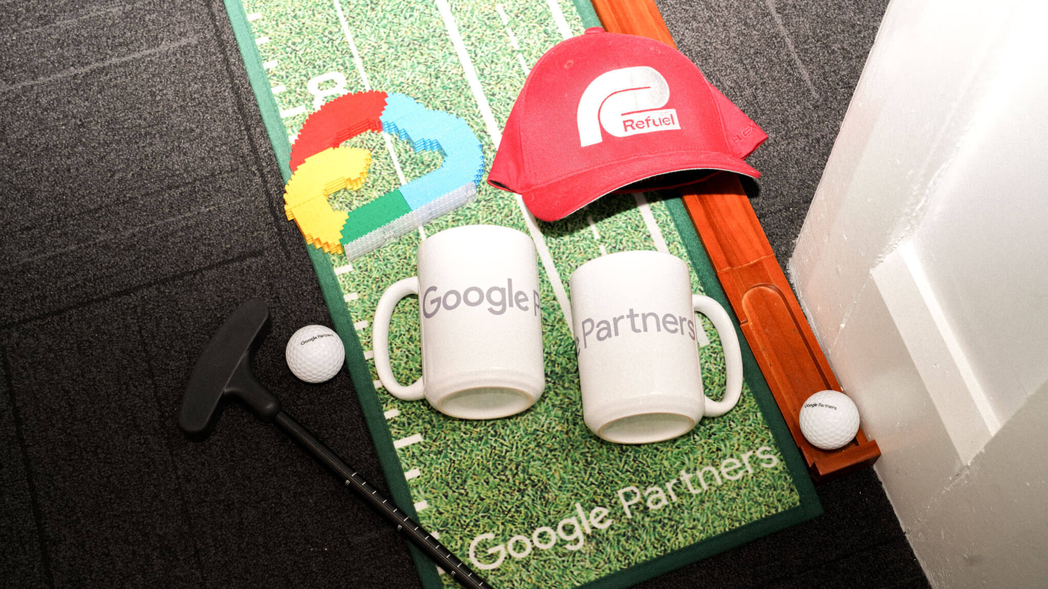 Google-branded golf merchandise, including a brick Google cloud logo, "Refuel" hat, "Google" and "Partners" mugs, putter, and balls, on a mini putting mat.
