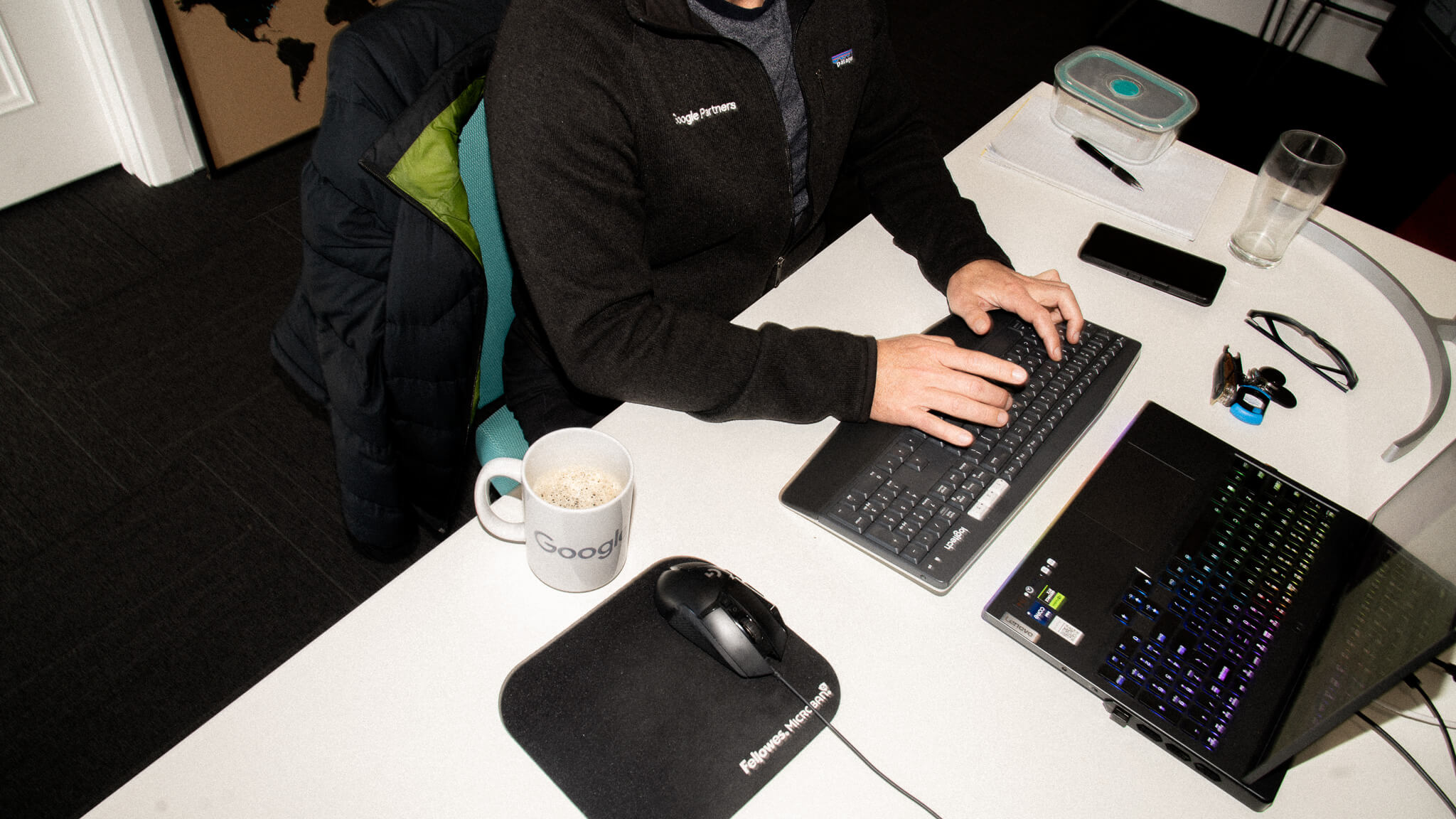 Viewed from above, a man in a Google Partners jacket types on a keyboard at his desk, with a Google mug and laptop nearby.
