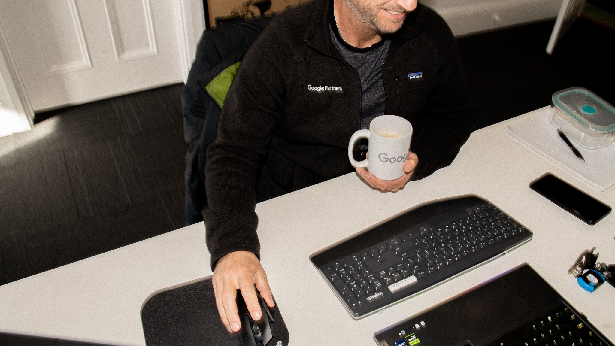 A man wearing a Google Partners jacket sits at his desk, holding a Google mug and using a computer mouse.