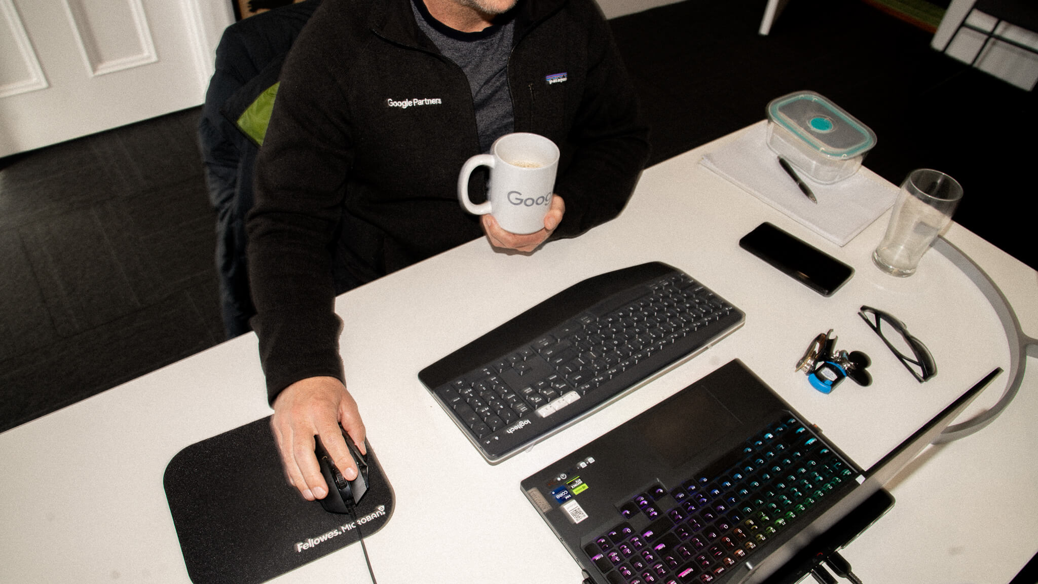 A man in a Google Partners jacket sits at his desk, holding a Google mug in one hand and using a computer mouse with the other.