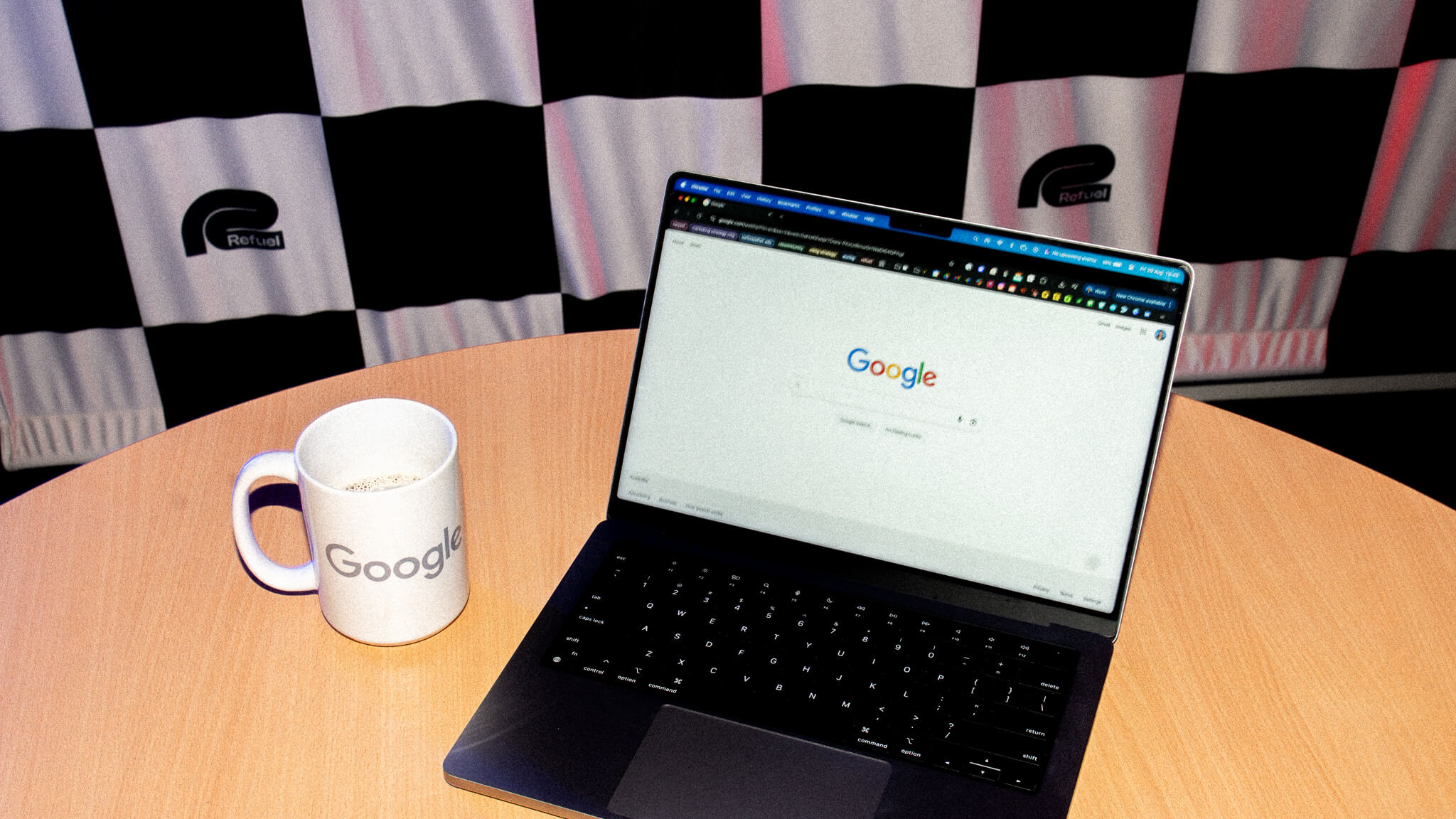 A laptop displaying the Google homepage and a Google mug sit on a table in front of a black and white checkered flag with the Refuel logo.