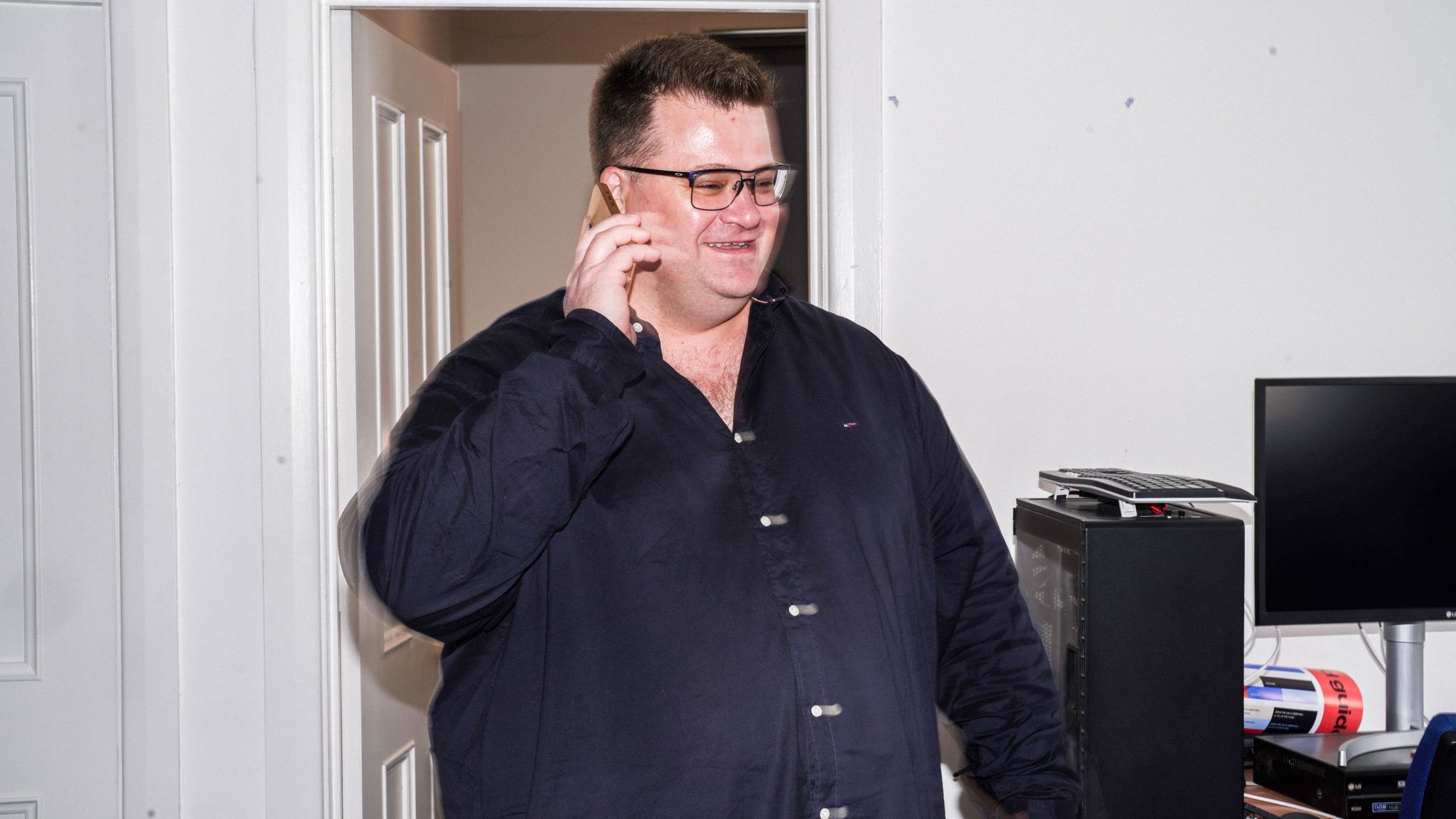 Person with glasses in dark button-up shirt smiling while talking on phone in doorway of office.