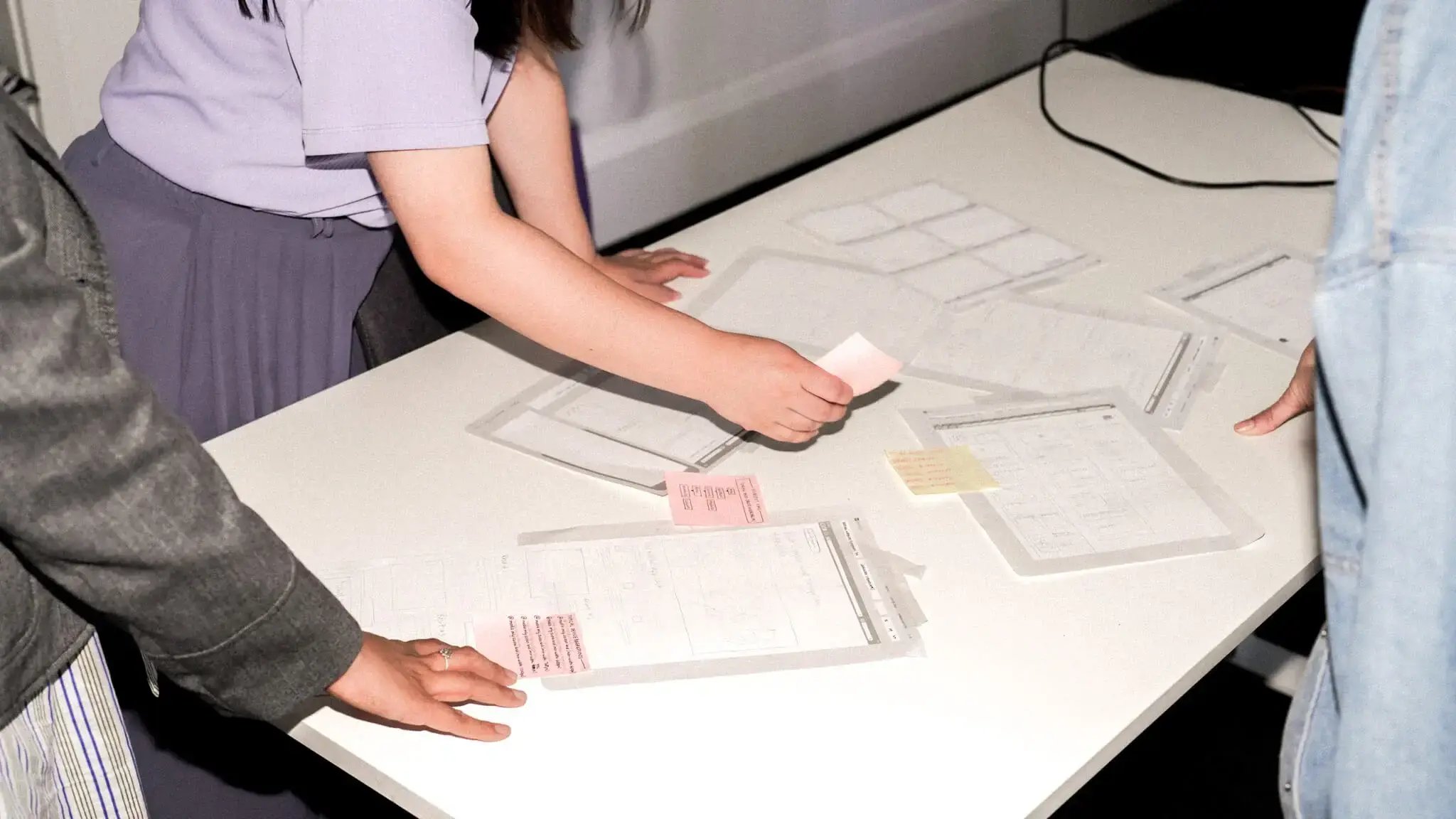 Hands arranging papers and sticky notes on a white table surface, showing a planning session in progress.