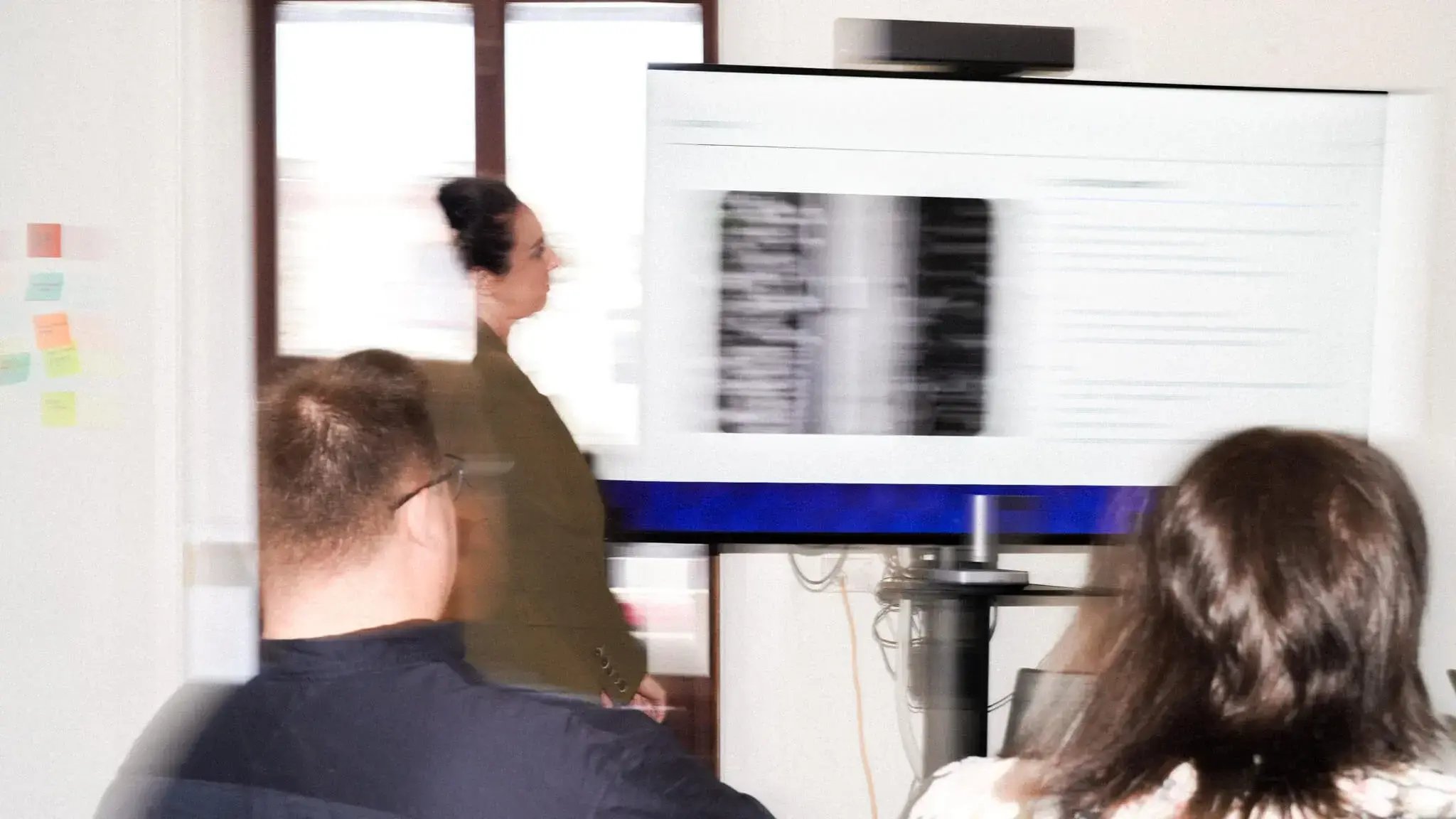 A dynamic, motion-blurred photo of a woman presenting to her team in a meeting room in front of a large screen.