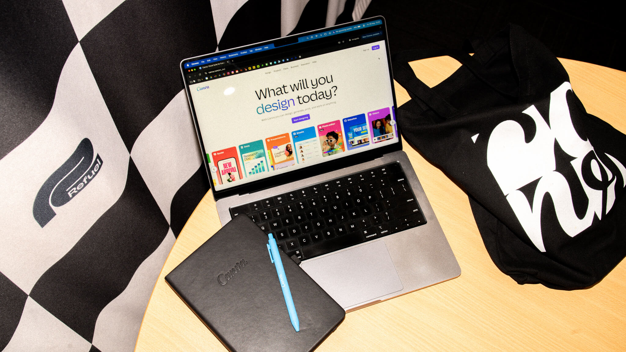 A laptop showing the Canva homepage sits on a table with a Canva notebook and tote bag, next to a Refuel-branded checkered flag.