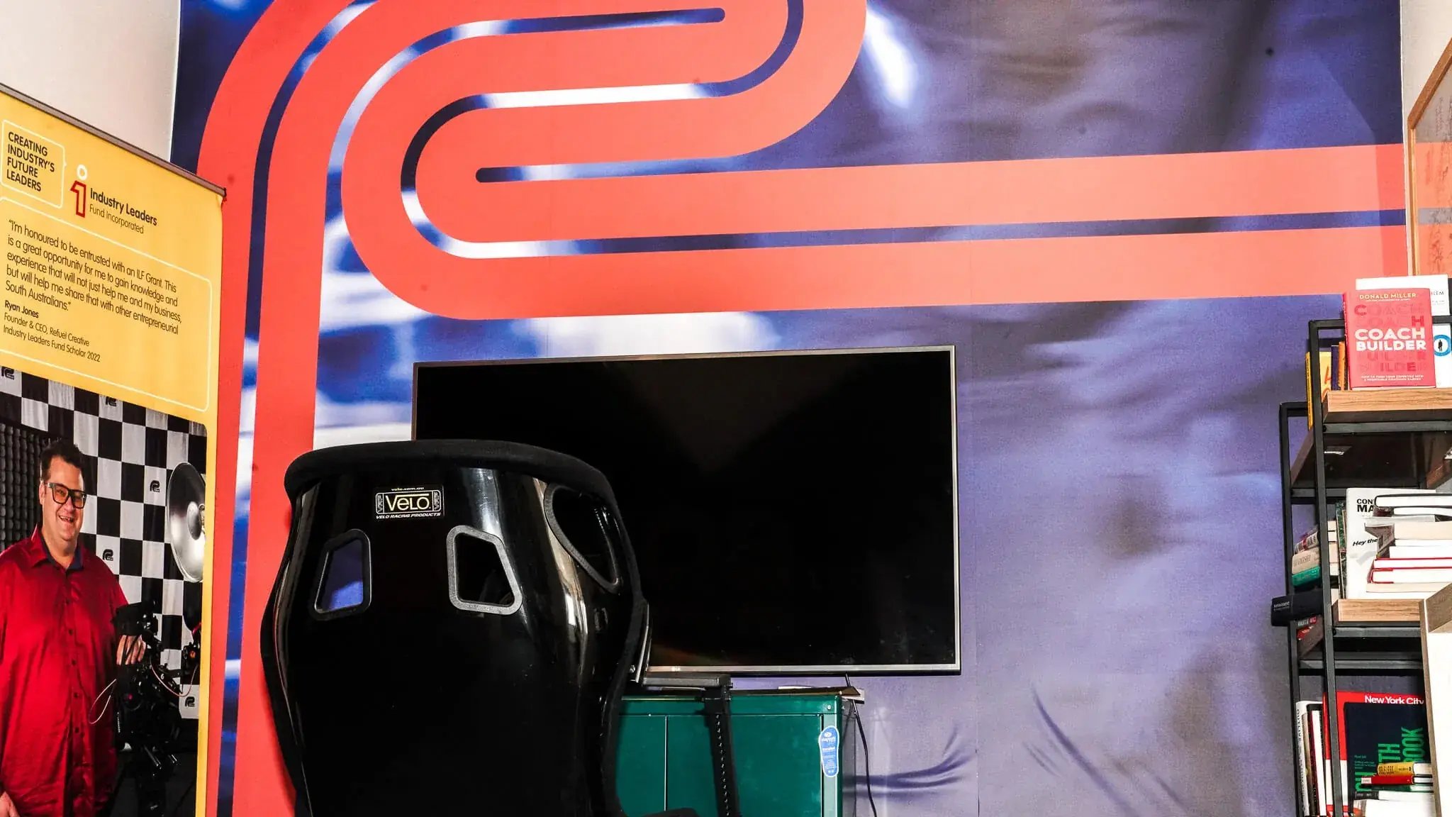 Racing-themed room with black gaming chair, large TV, checkered wall pattern, and curved red/blue racing stripe ceiling design.