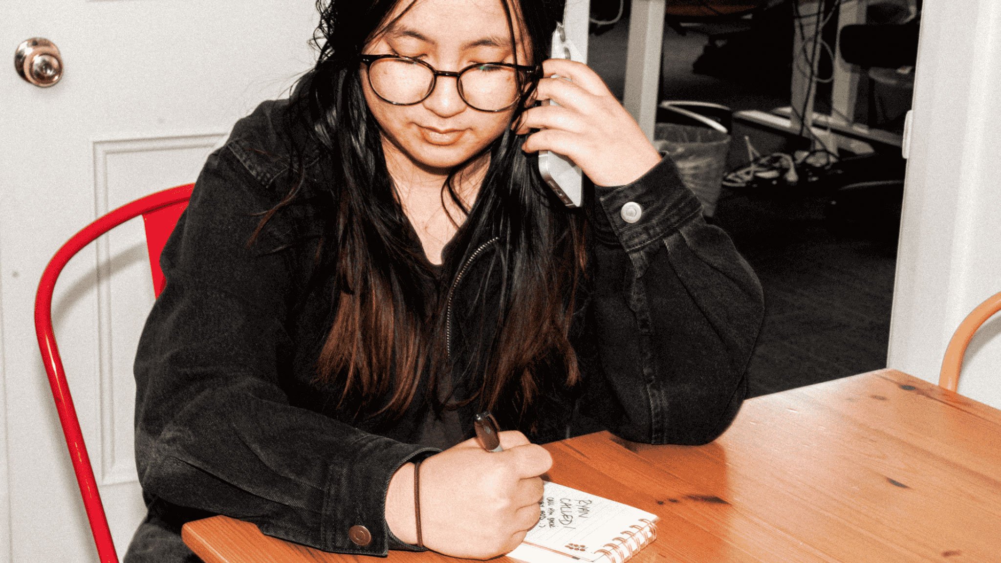 Person with long dark hair and round glasses in black jacket on phone while writing in notebook at wooden desk.