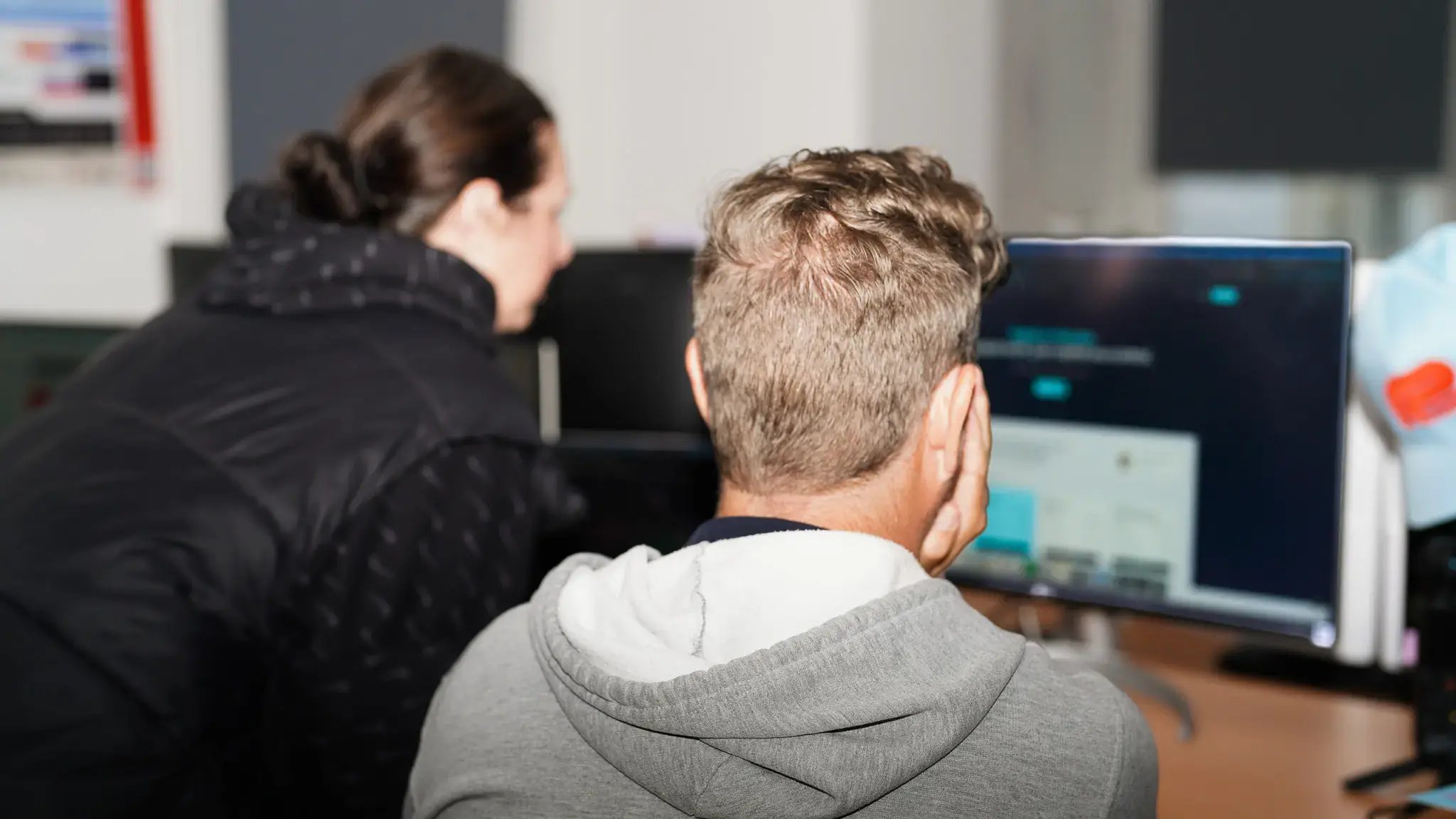 A rear view of two colleagues, a woman and a man, sitting side-by-side at a desk. They are collaborating and looking intently at a computer screen, which is slightly out of focus.