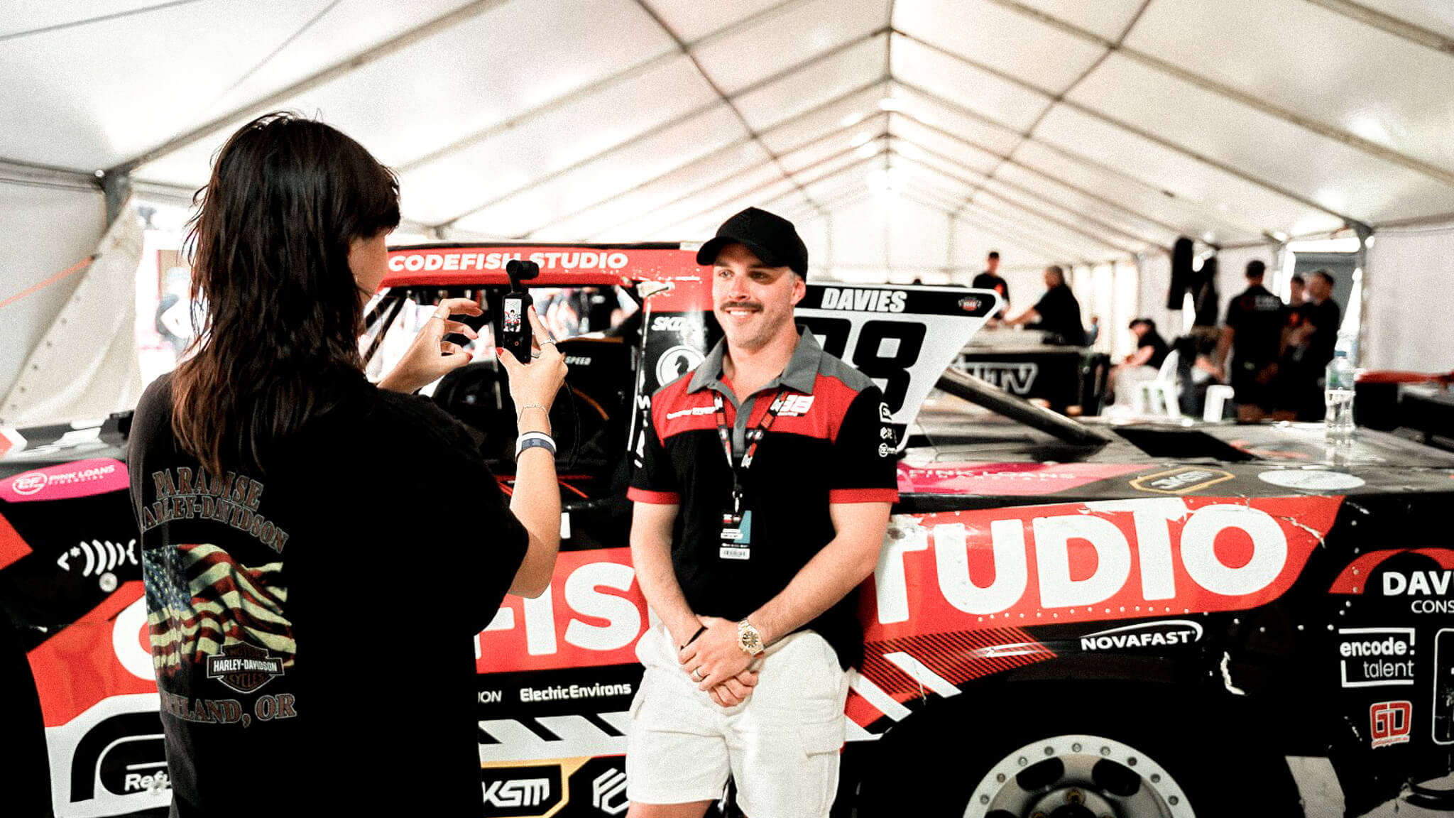 Someone in a black t-shirt taking a photo of a race car driver standing next to a red and black Codefish Studio super truck in a white tent paddock area.