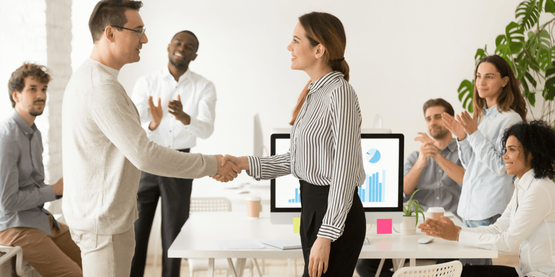 man and woman shaking hands in an office environment surrounded by team members
