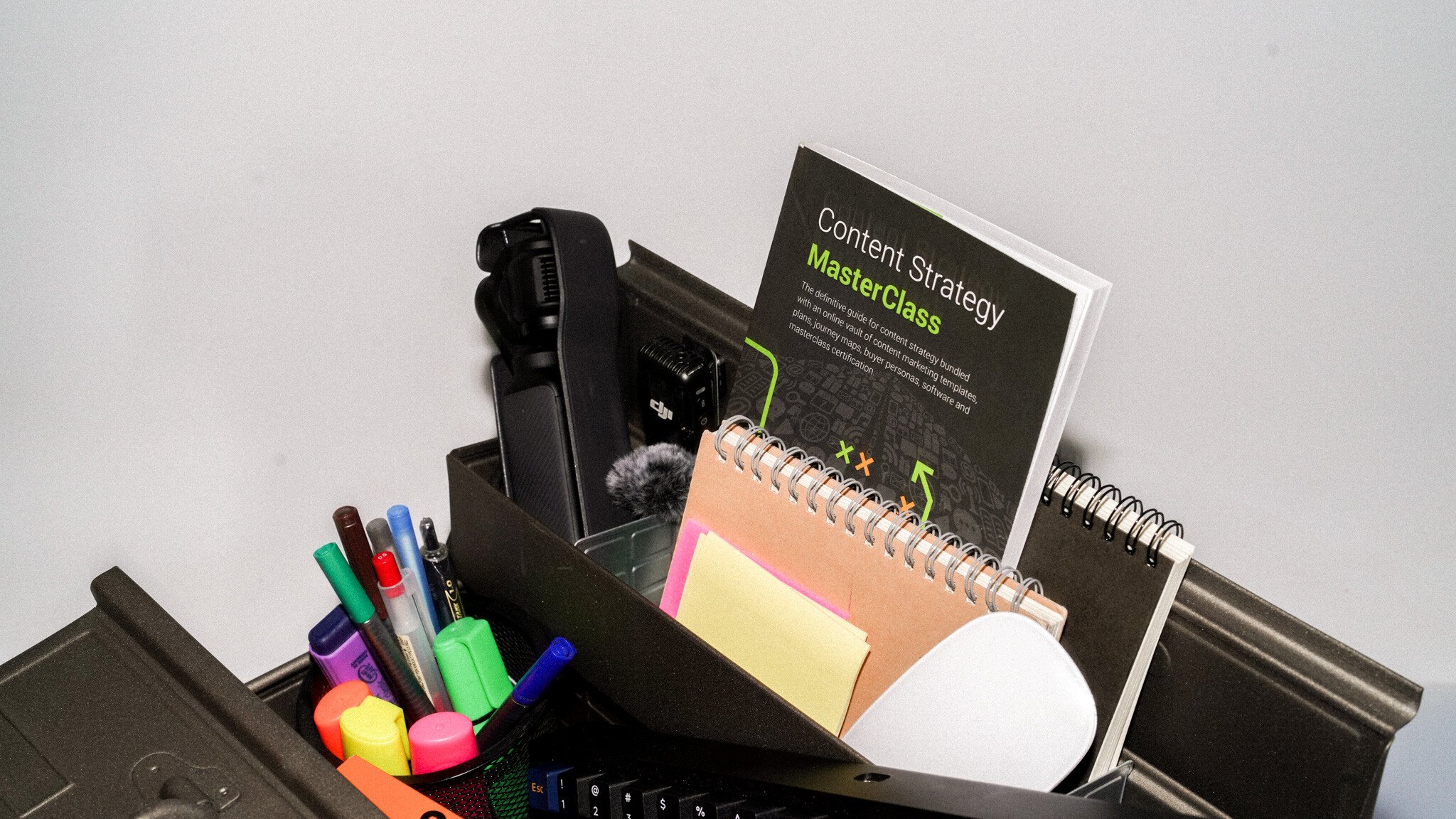 A tool box containing colourful textas and highlighters, a spiral notepad, sticky notes, and a book titled 'Content Strategy MasterClass' with what appears to be strategic diagrams on its dark cover. The items are arranged against a light grey background.