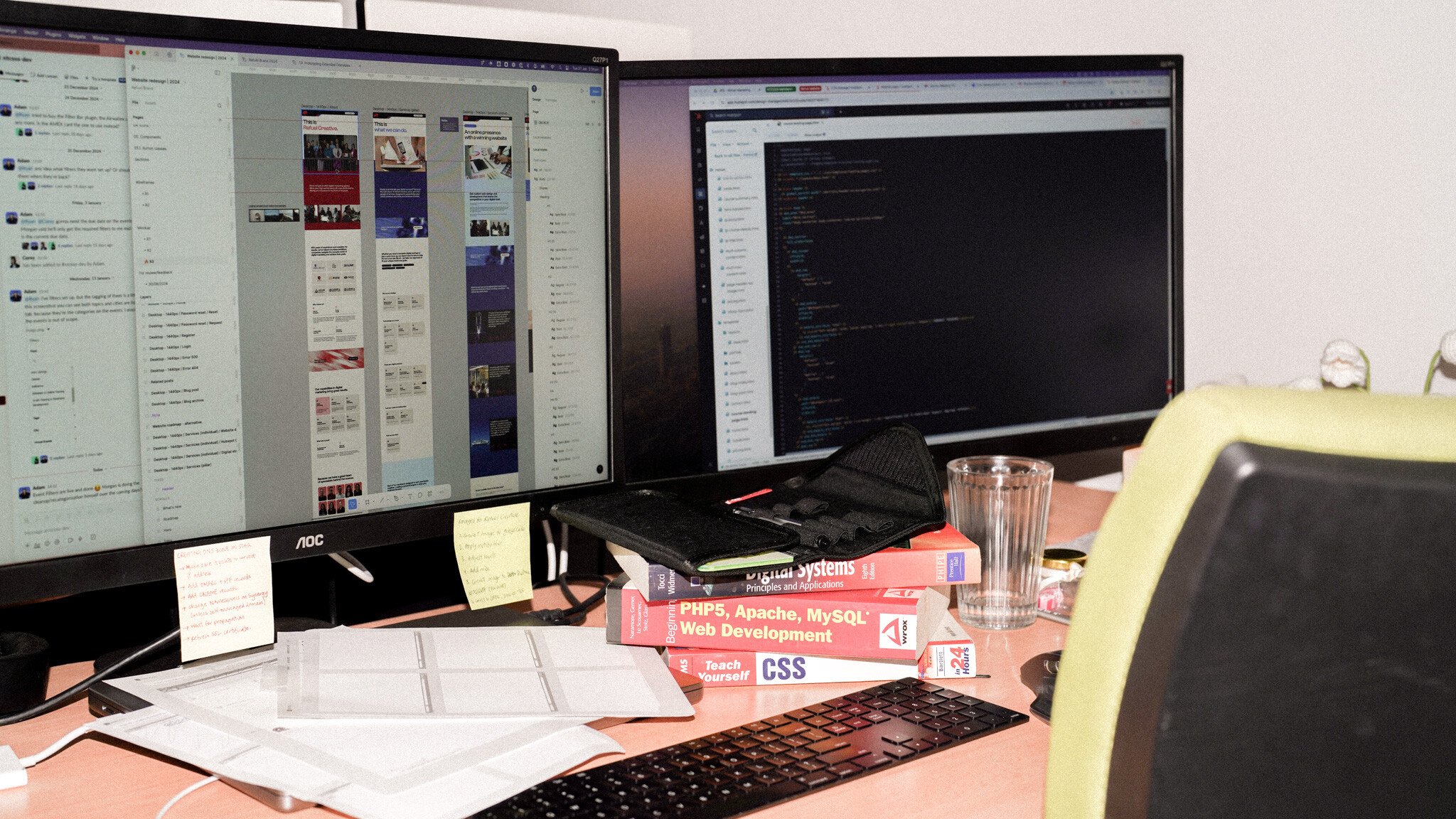 A developer's workspace with dual monitors showing website layouts and code editor with dark theme. Programming books including 'PHP5, Apache, MySQL' and 'CSS' are stacked on the desk alongside a keyboard, sticky notes, and a water glass.