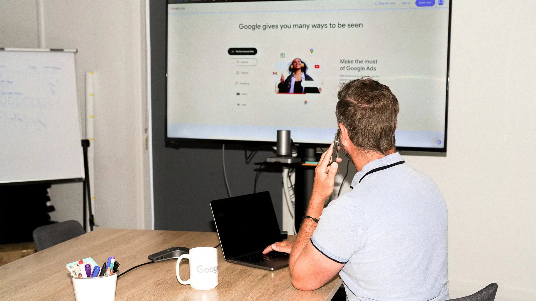 A man in a light blue polo sits at a desk, talking on a phone and using a laptop. A large screen behind him displays a Google Ads presentation. A Google-branded mug sits nearby.