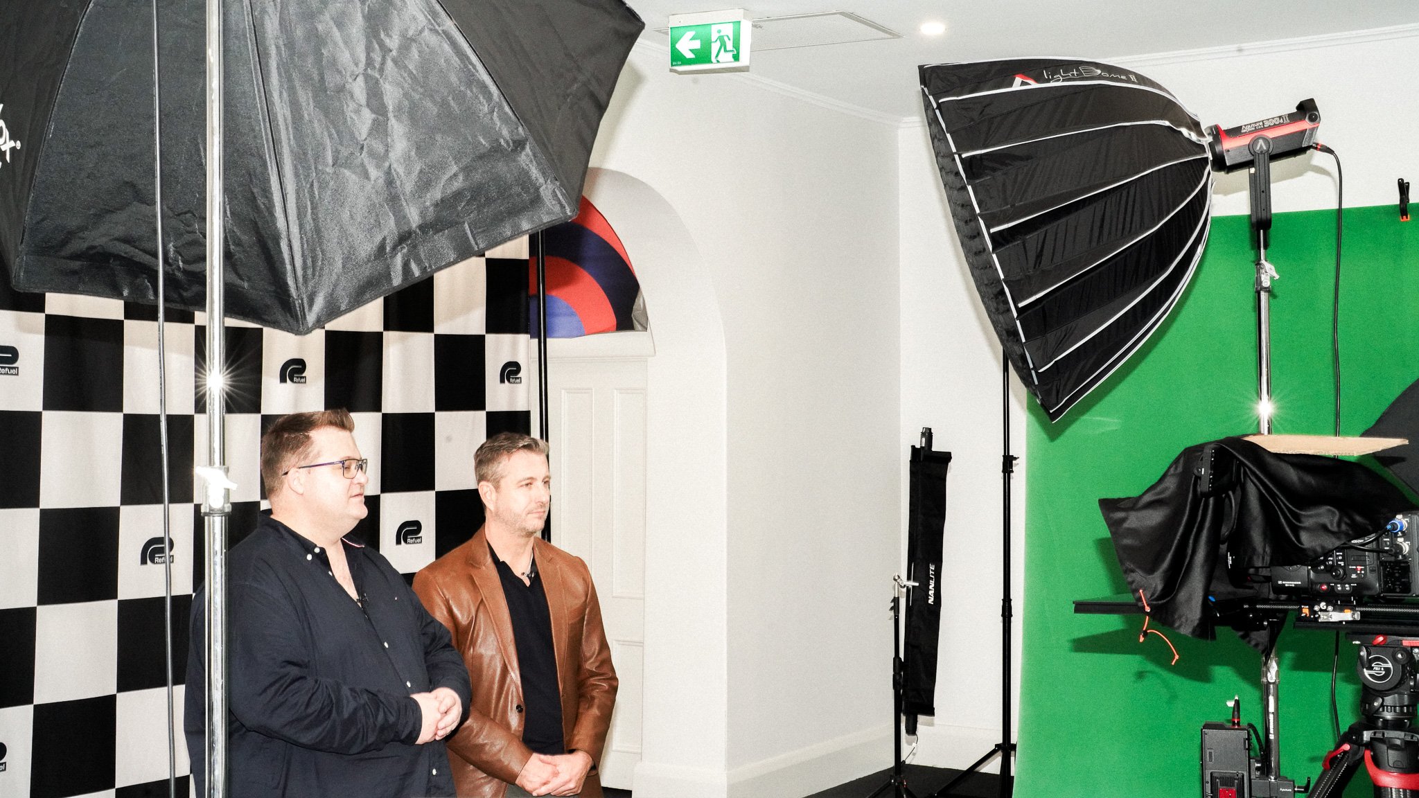 Professional photo/video studio with two men, checkered backdrop, green screen, and professional lighting equipment including softboxes and umbrellas.