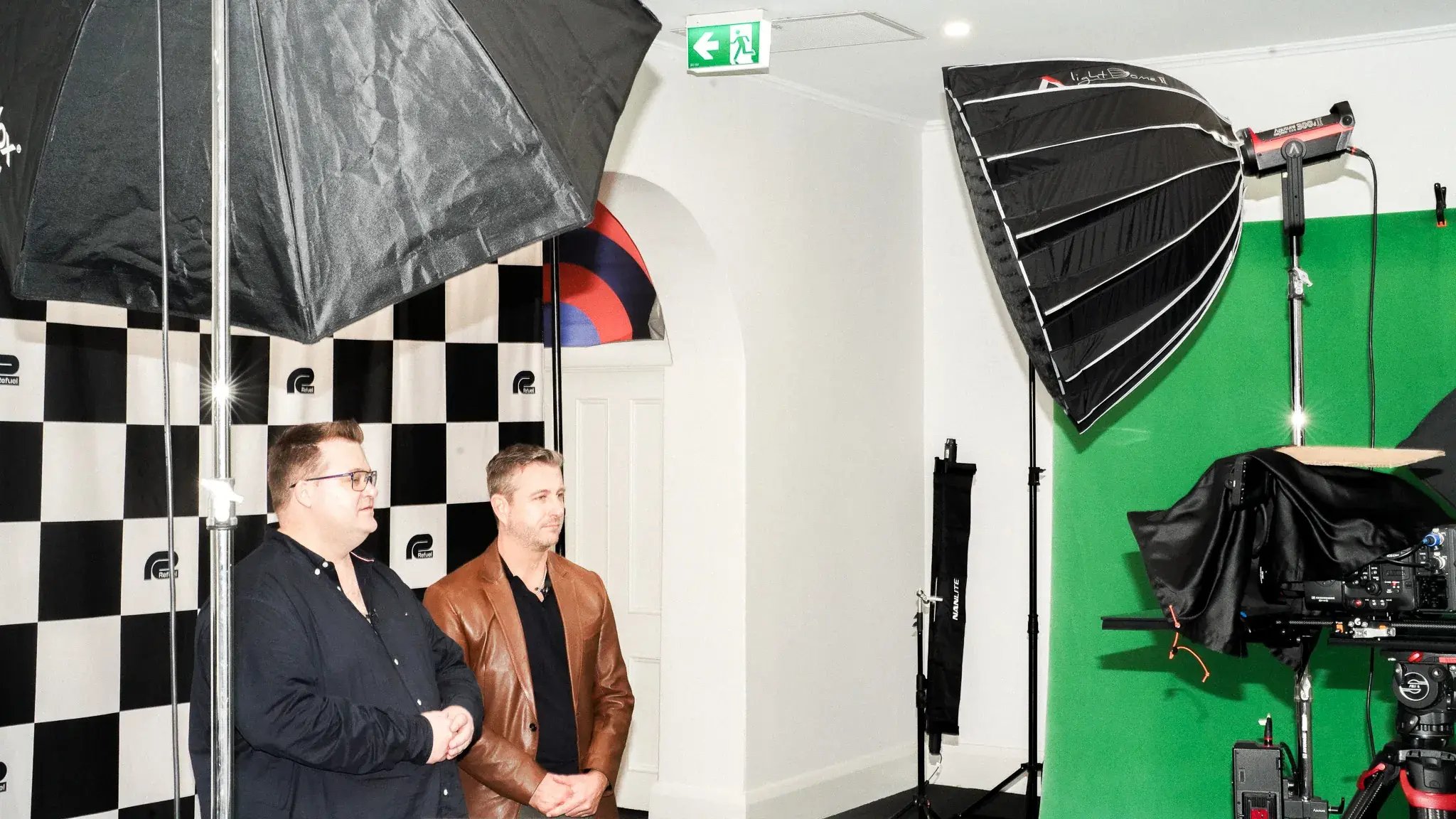 Professional photo/video studio with two men, checkered backdrop, green screen, and professional lighting equipment including softboxes and umbrellas.