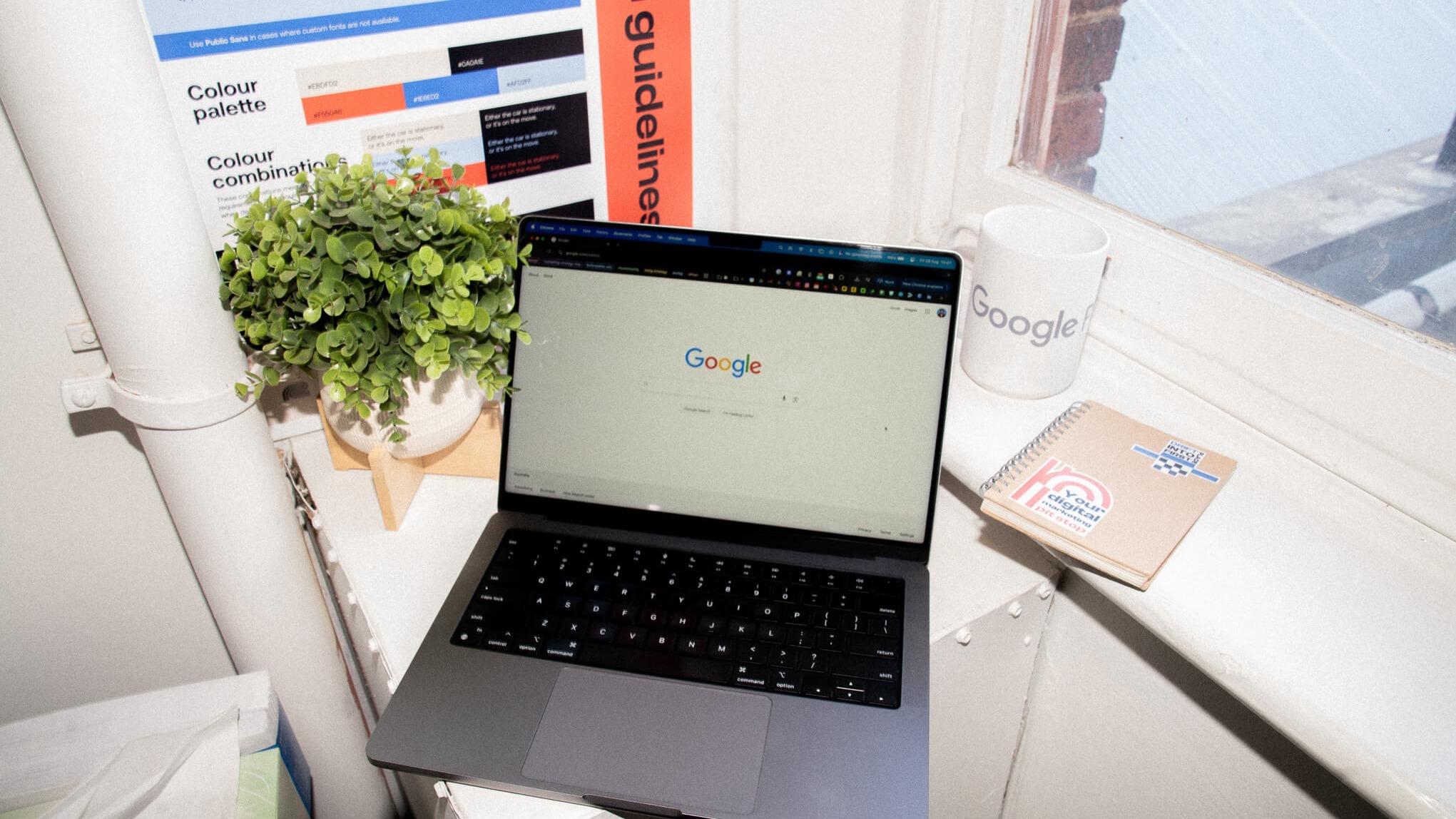 A laptop showing the Google search page on a windowsill, next to a potted plant, a Google mug, and a poster of brand colour guidelines.
