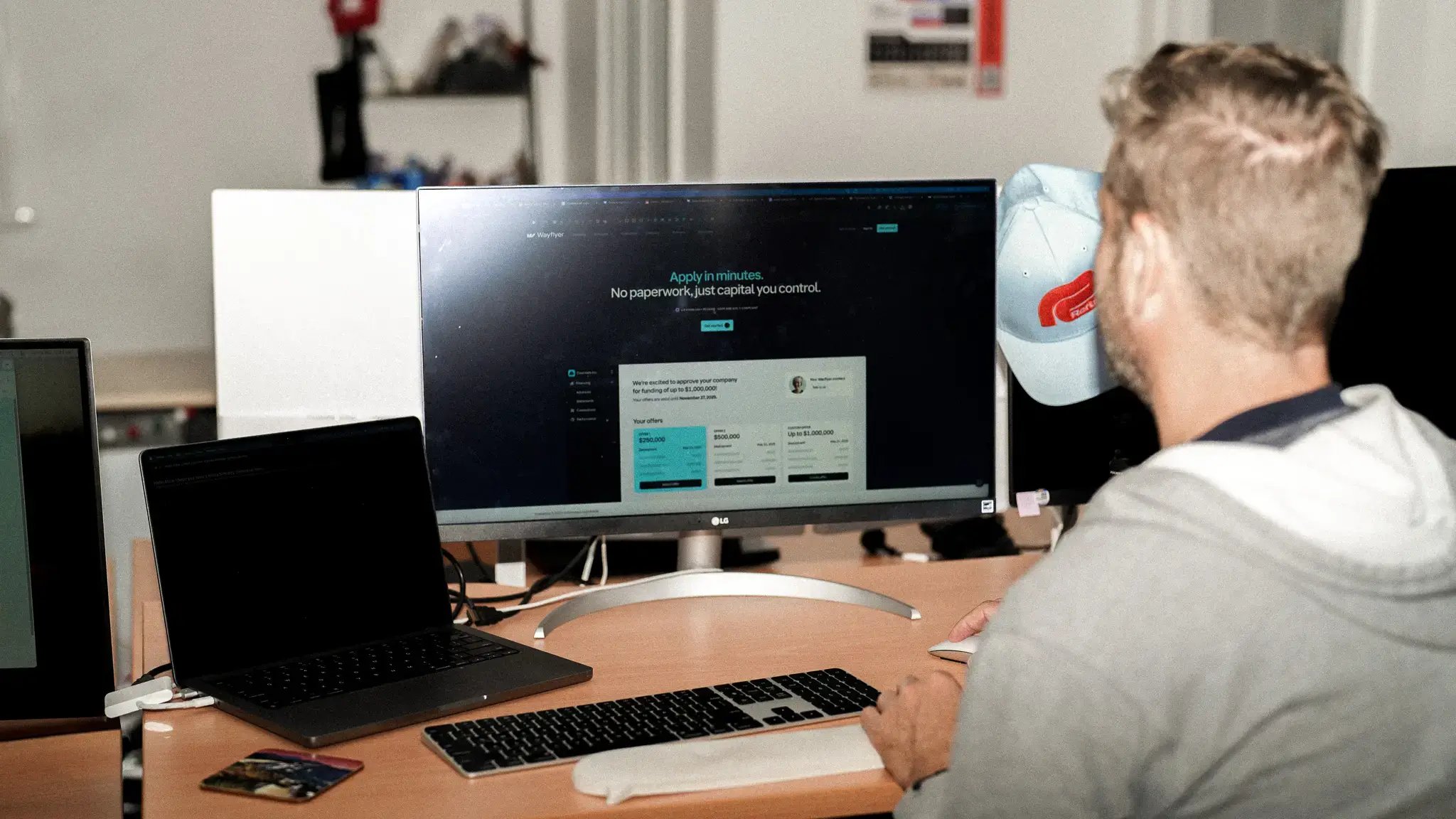 A wide-angle shot of a person working at a desk, viewed from behind. They are using a mouse and keyboard while viewing the Wayflyer website on a large LG monitor. The workspace includes an open laptop to the left and a cluttered office background.