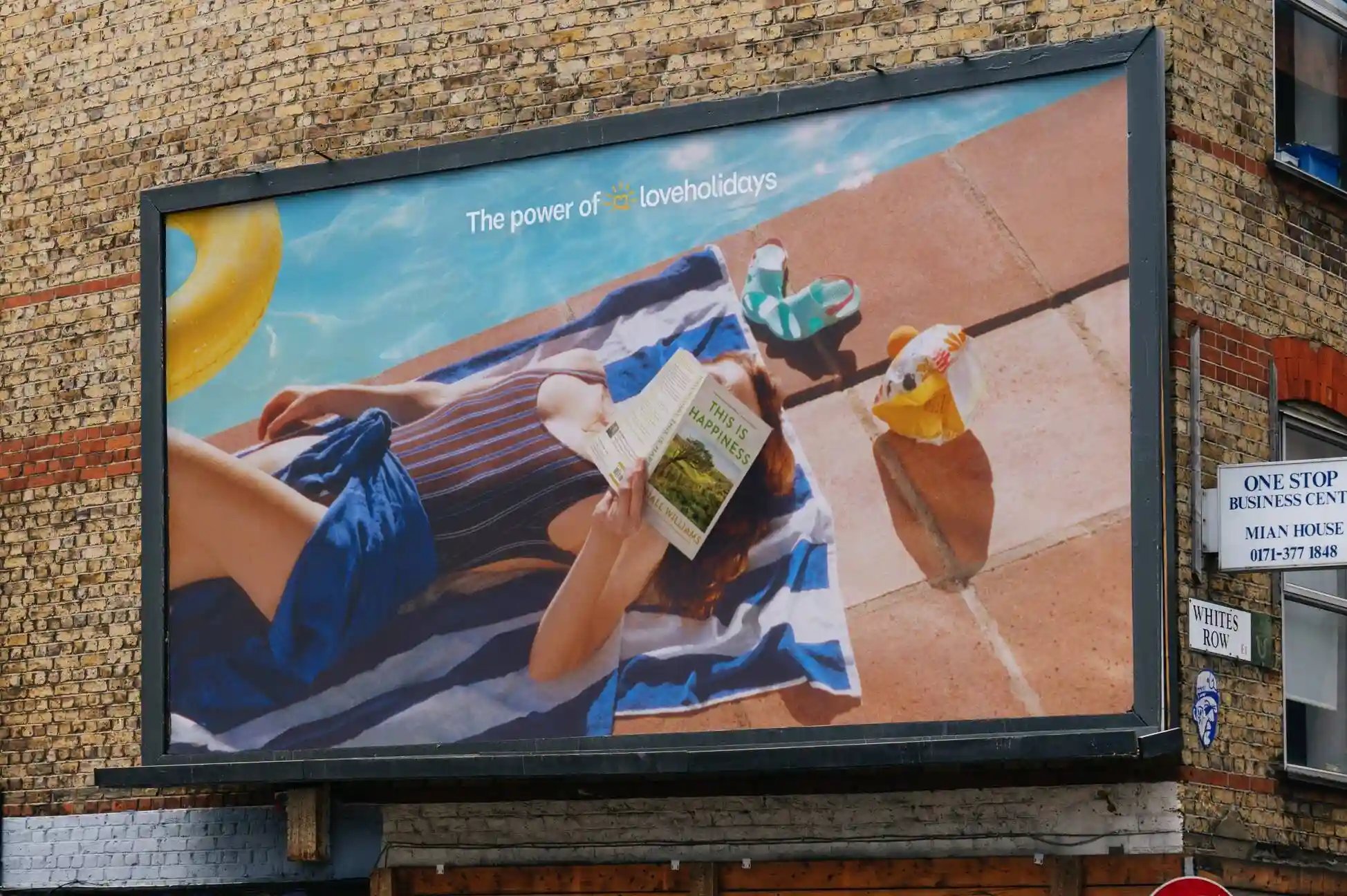 loveholidays outdoor advertising campaign using visual storytelling and 'in media res' techniques, featuring a sunbather hiding behind a book titled 'This is Happiness' to evoke relaxation.