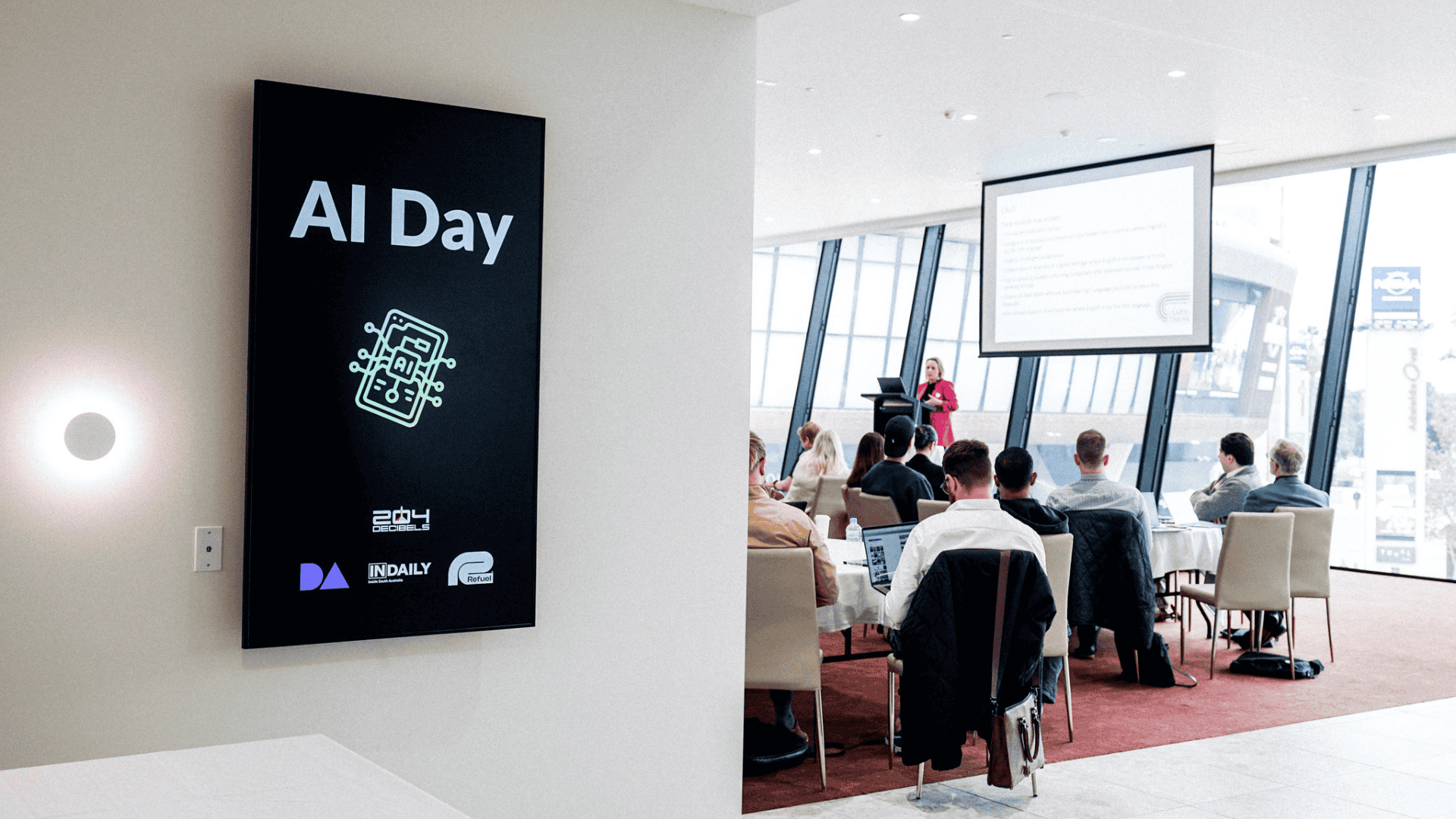 "AI Day" conference sign on wall beside meeting room with attendees watching presenter at projection screen.