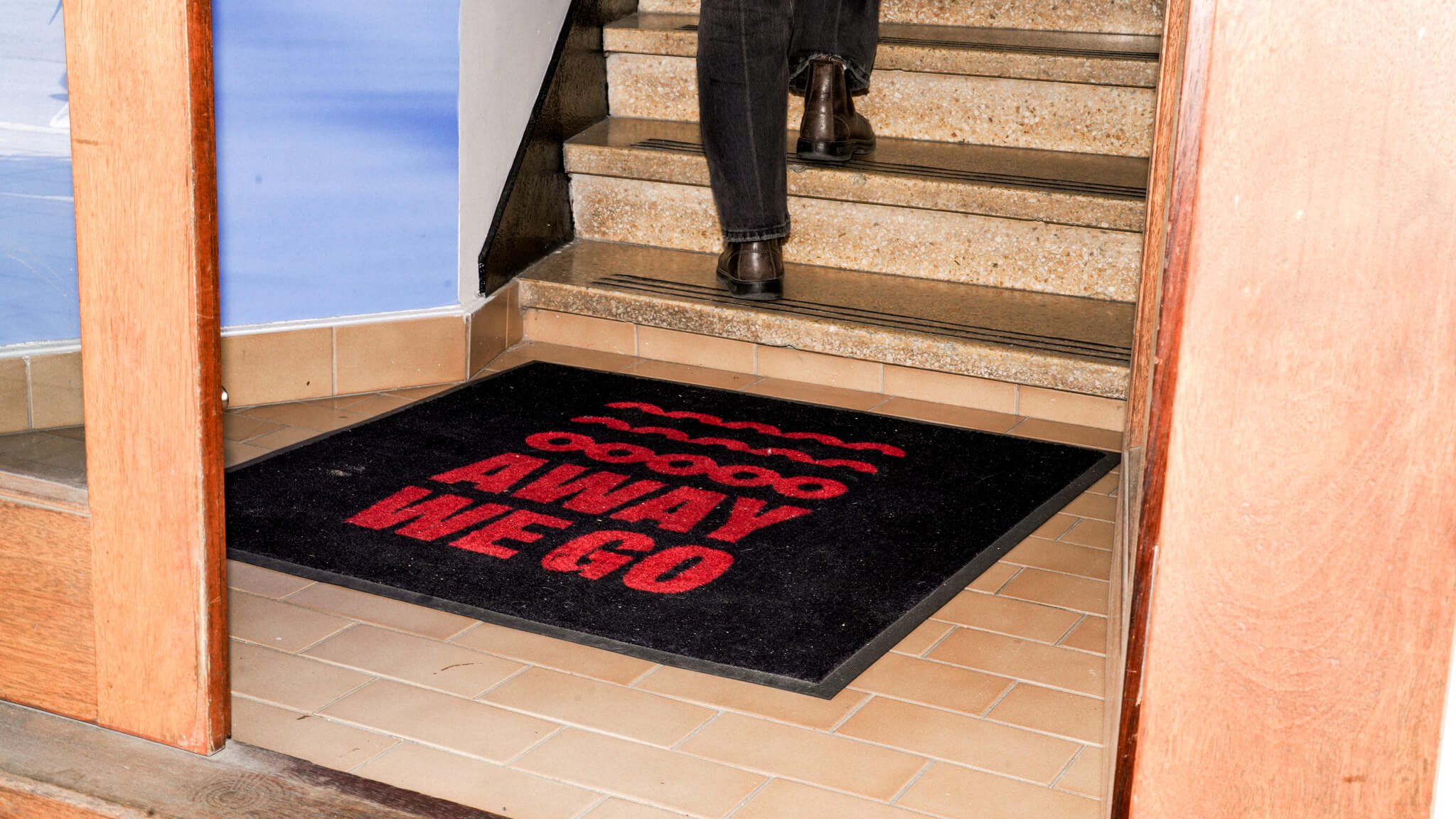 A black doormat with red text reading 'Away we go' at the bottom of a tiled staircase. Someone's feet in black pants and boots are visible on the stairs.