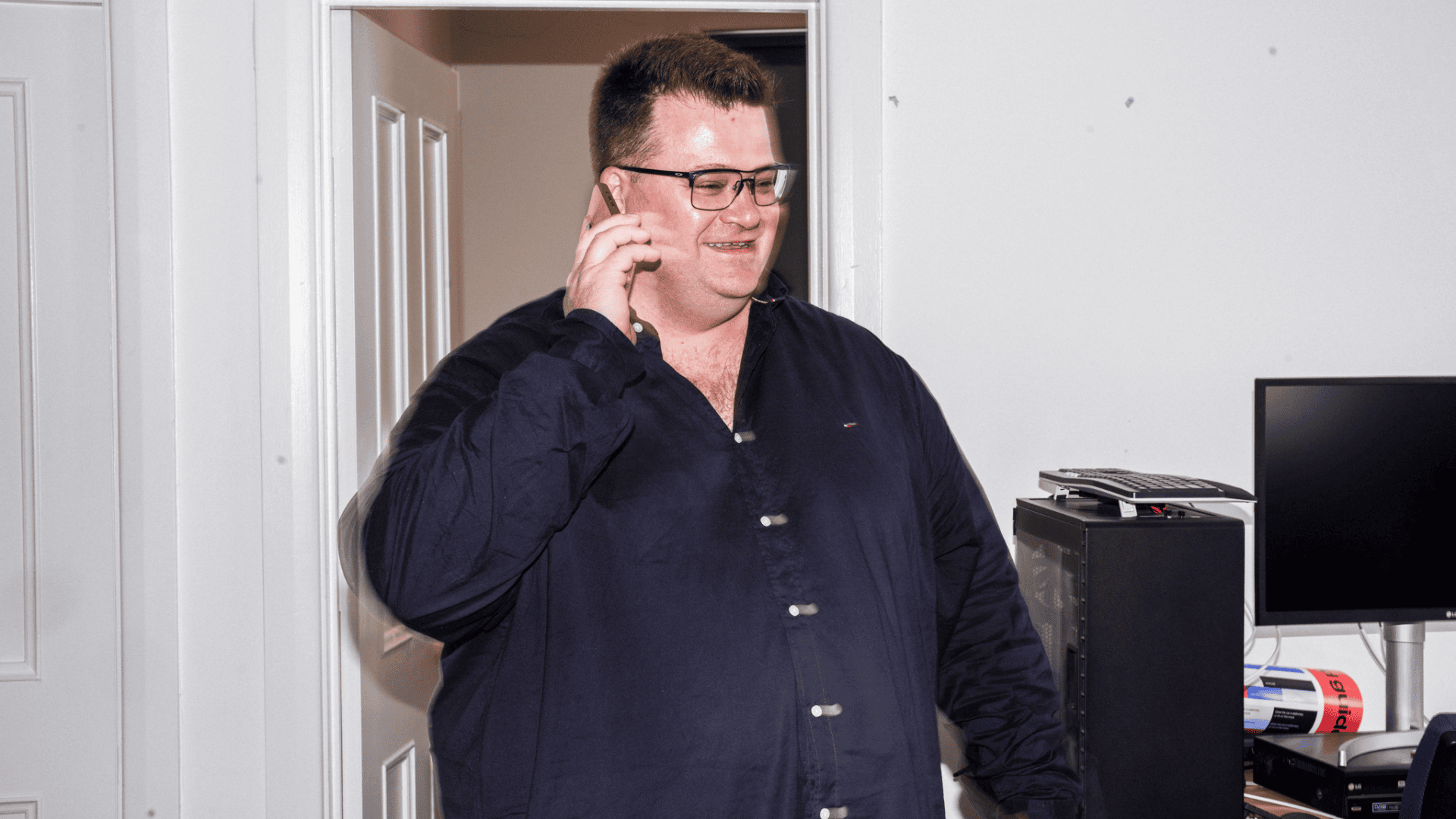 Person with glasses in dark button-up shirt smiling while talking on phone in doorway of office.