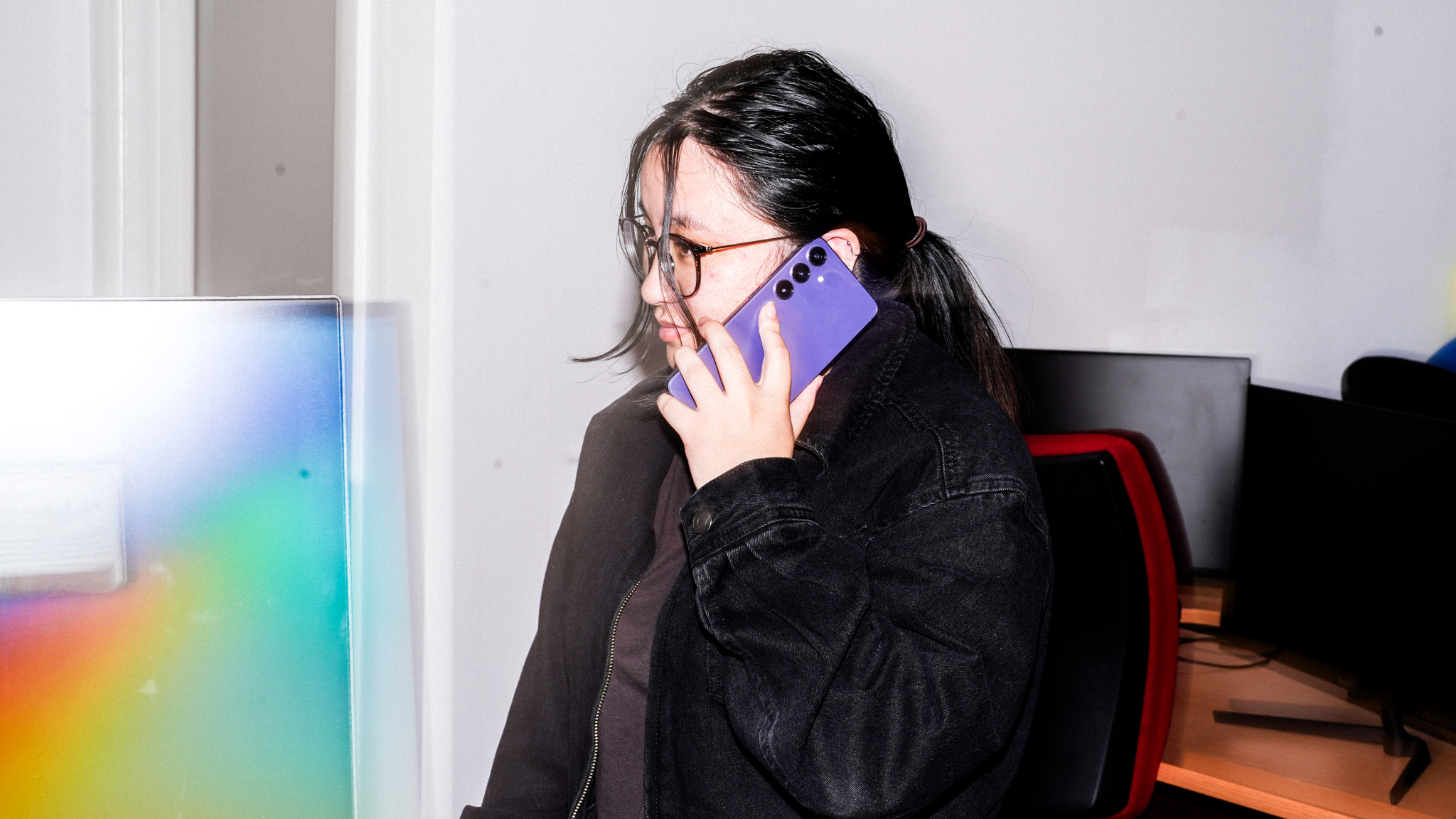 Person with glasses in black jacket on call with purple phone, standing near colourful screen in indoor setting.