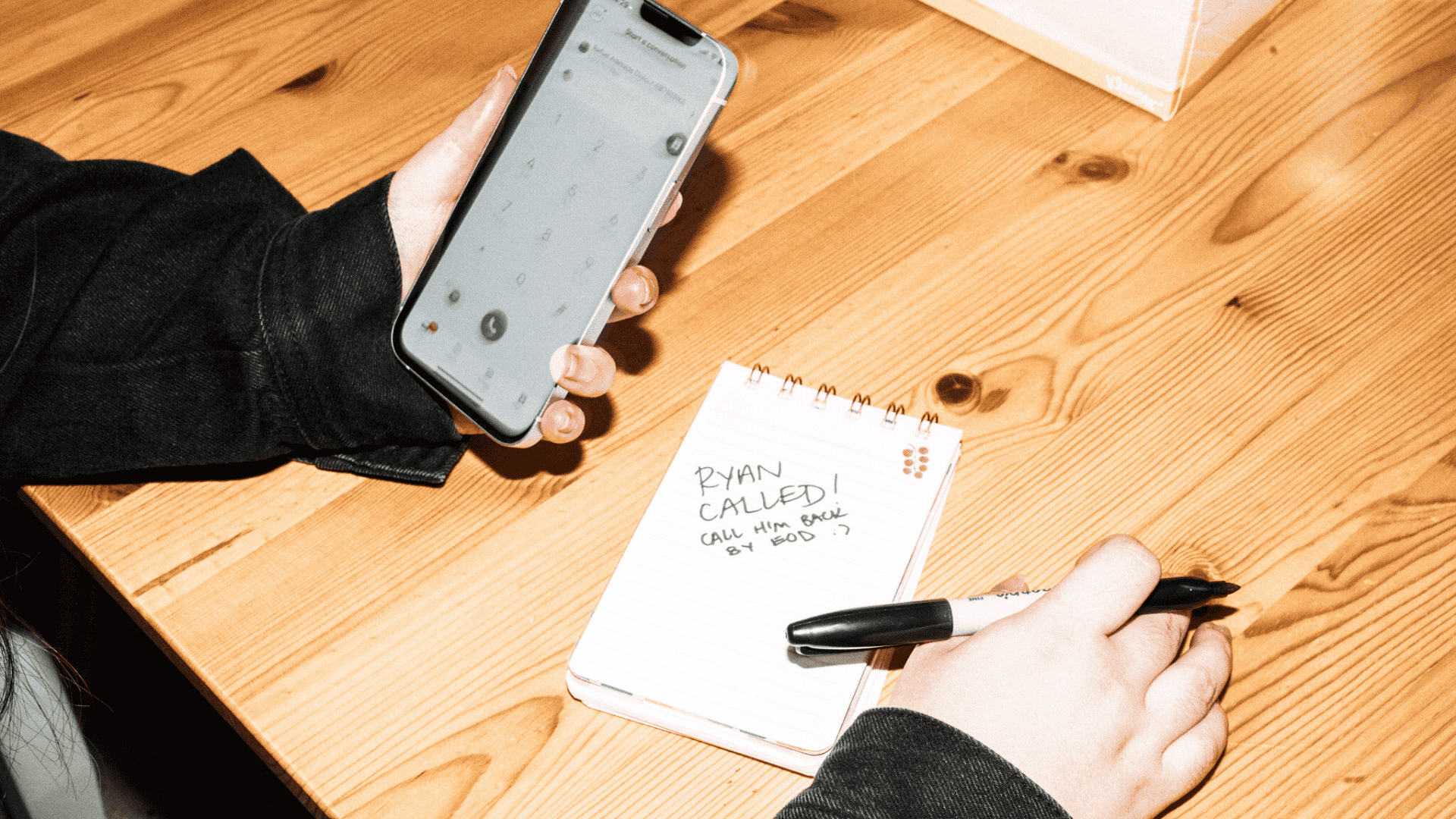 Person in black sleeves holding phone while writing "Ryan called" note on spiral notebook on wooden table.
