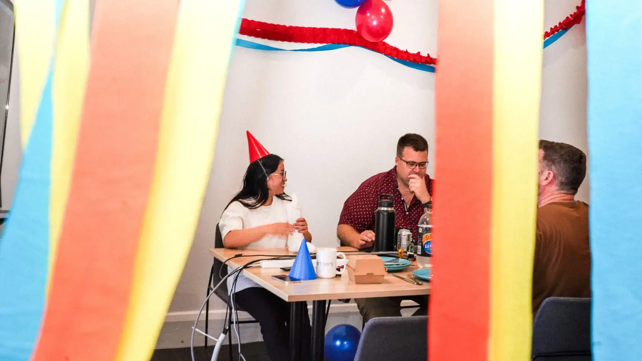 Office celebration with colourful streamers, party hats, and coworkers gathered around table.