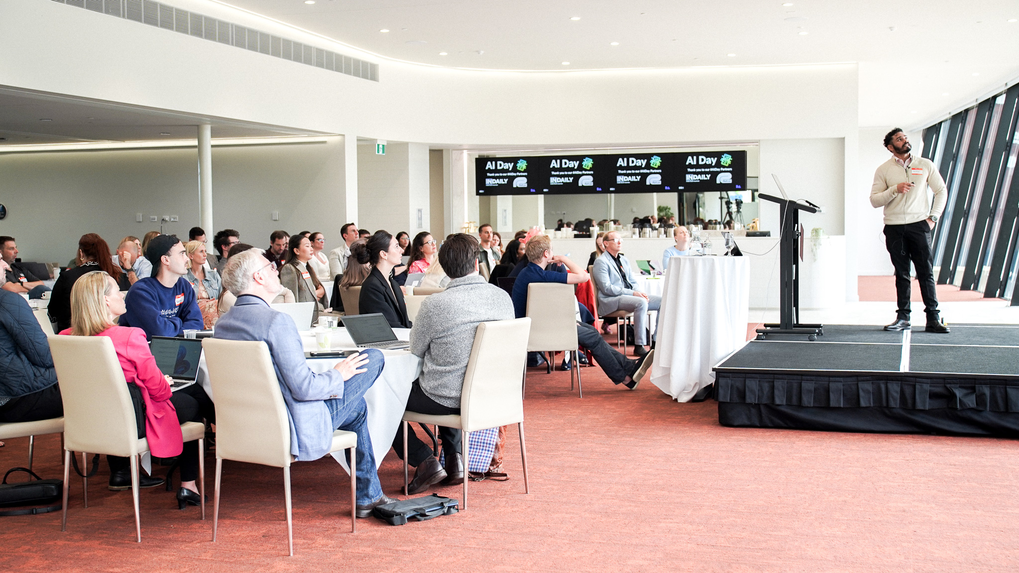 Speaker presenting to seated audience at AI Day conference in modern venue with digital screens and professional staging setup.