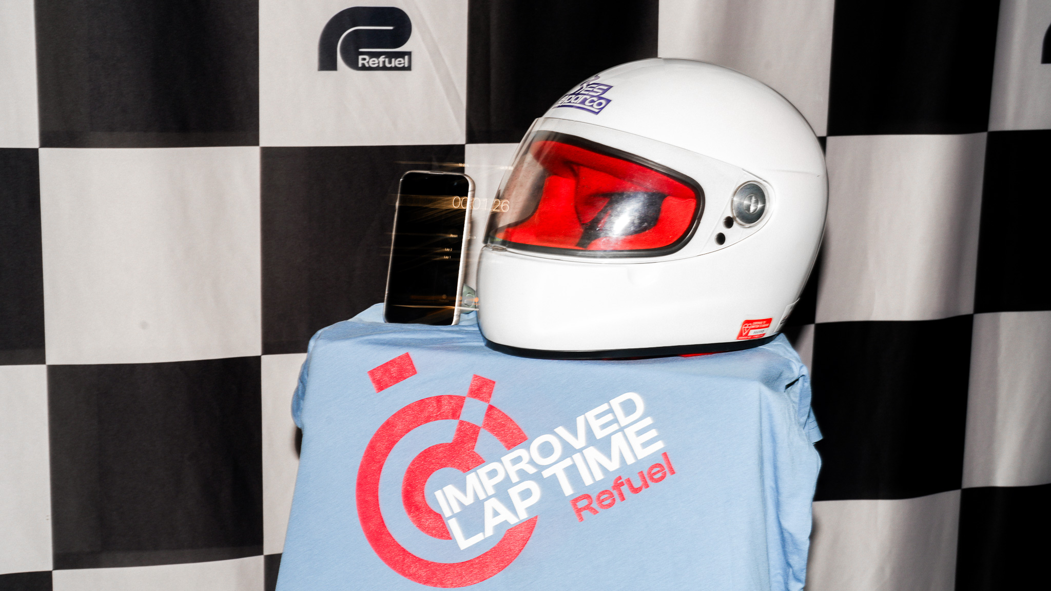 White racing helmet with red visor and phone showing timer on checkered flag backdrop with Refuel branding.