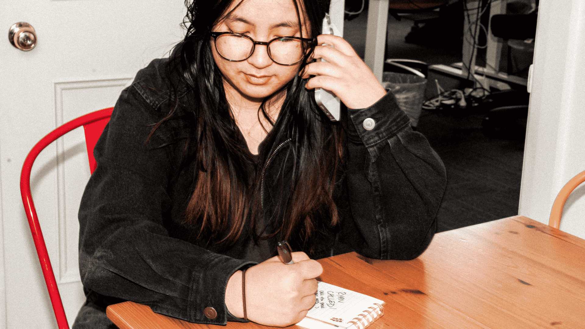 Person with long dark hair and round glasses in black jacket on phone while writing in notebook at wooden desk.