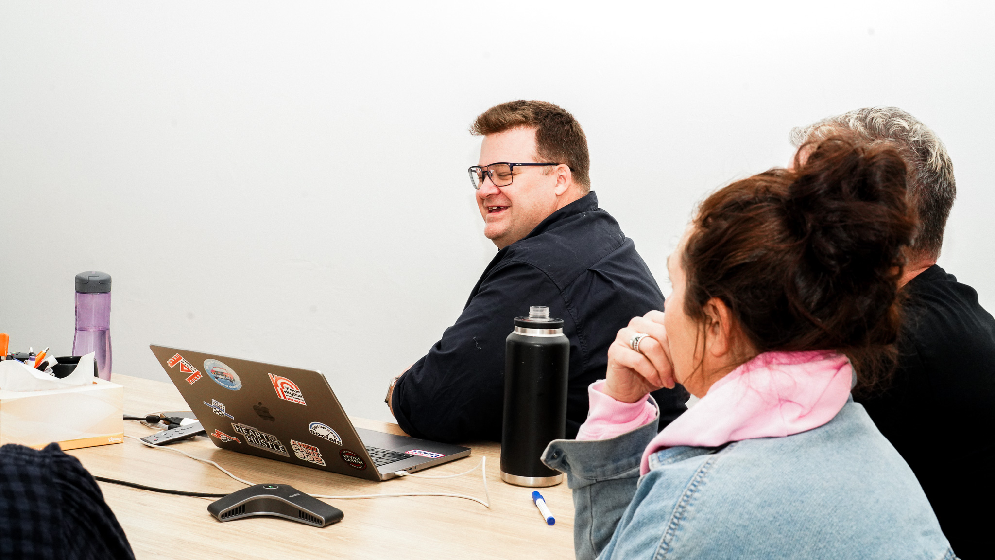 Team meeting with man in glasses at laptop with stickers, woman in pink hoodie and denim jacket, and another person at wooden table with water bottle and mouse.