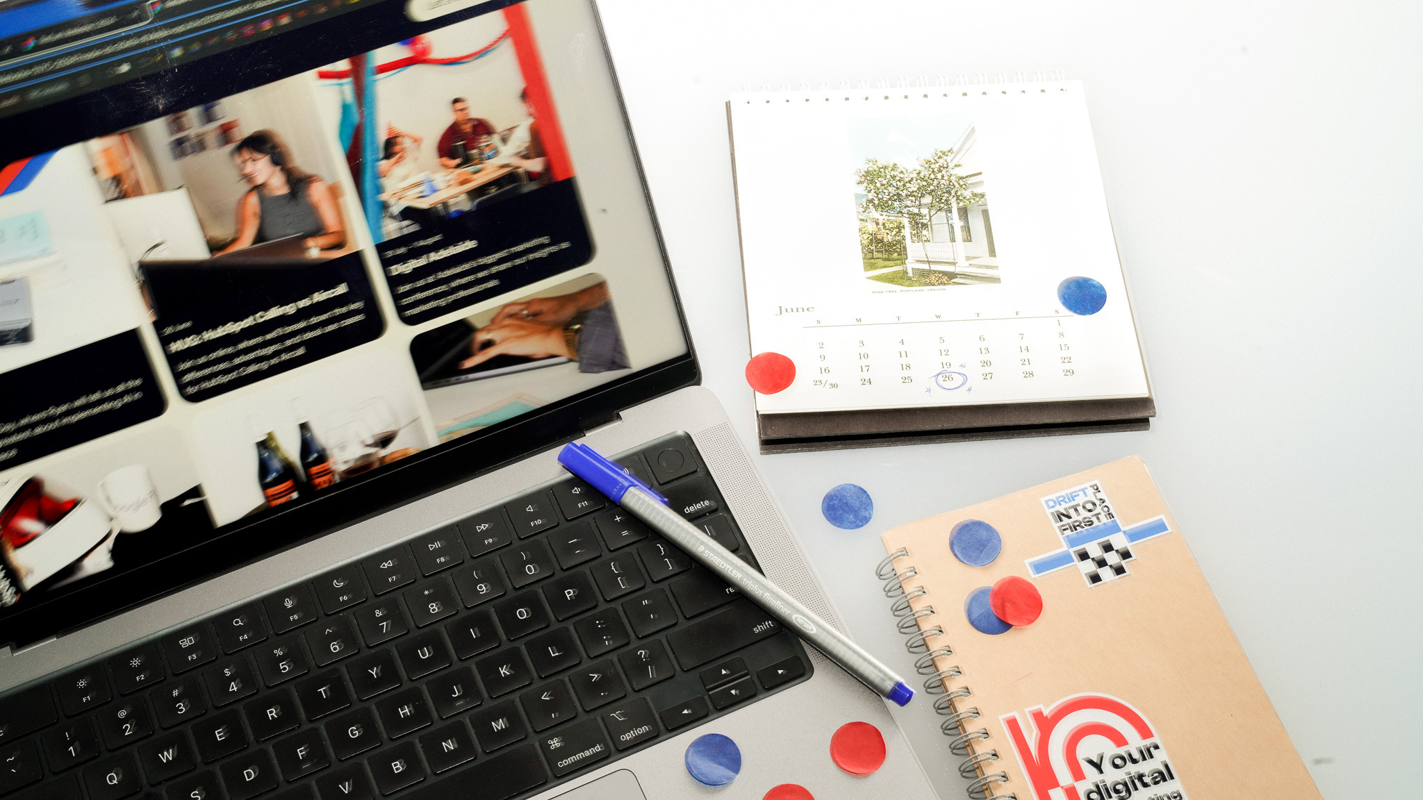 Laptop with video thumbnails, June calendar, notebook with red and blue confetti, and blue pen on white desk.