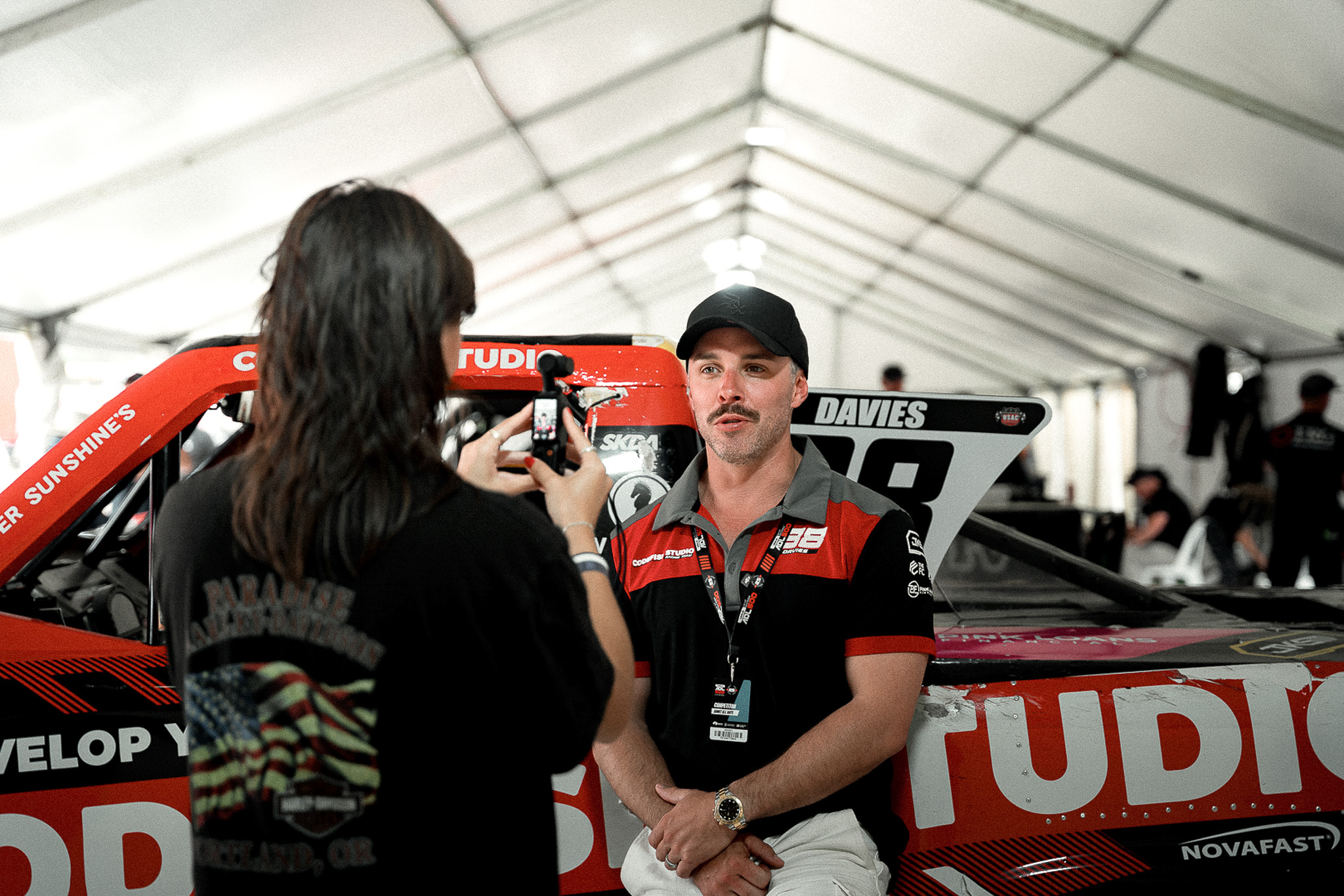 Someone in a black t-shirt taking a photo of a race car driver standing next to a red and black super truck in a white tent paddock area.