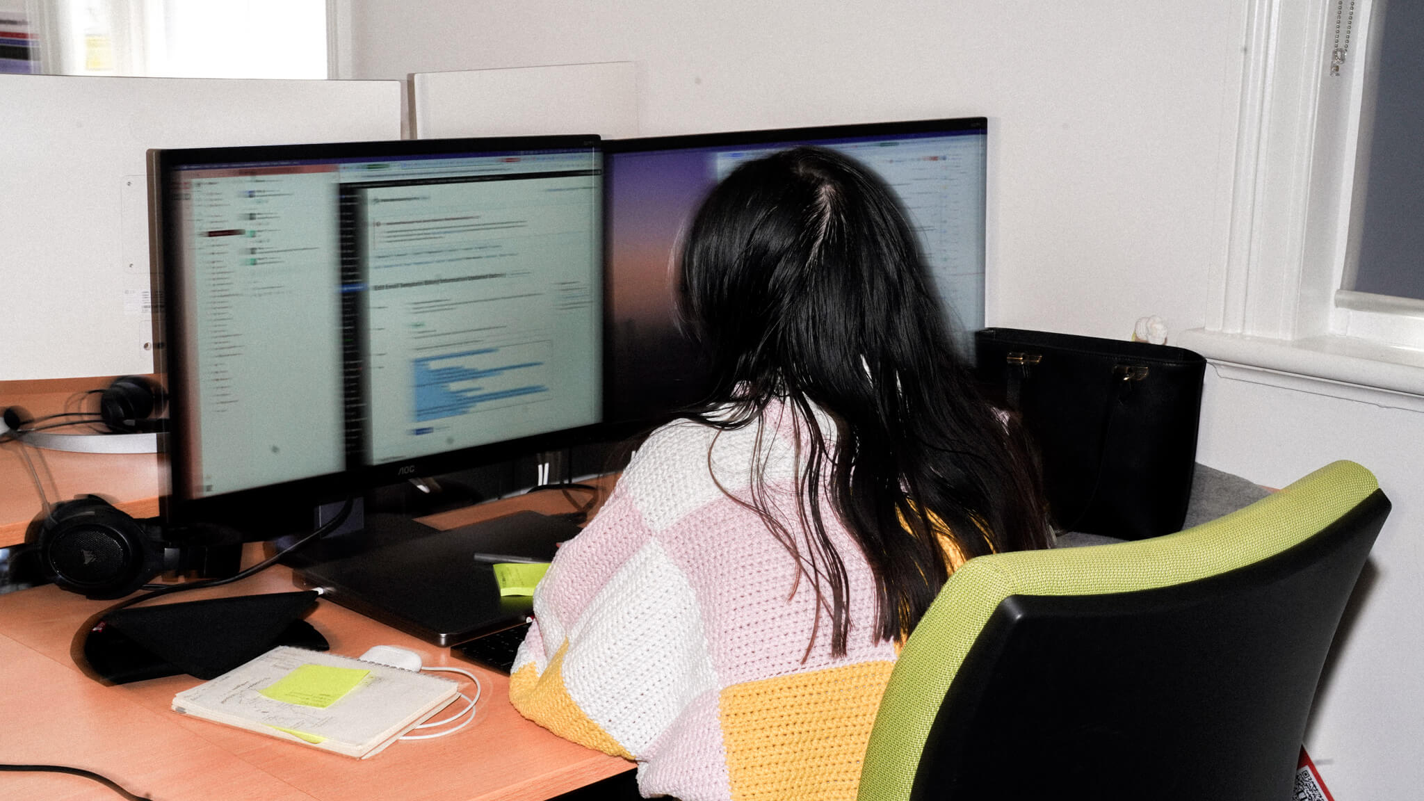 Person with long dark hair in colourful knit sweater working at dual-monitor setup with code visible on screens, seated in green office chair.