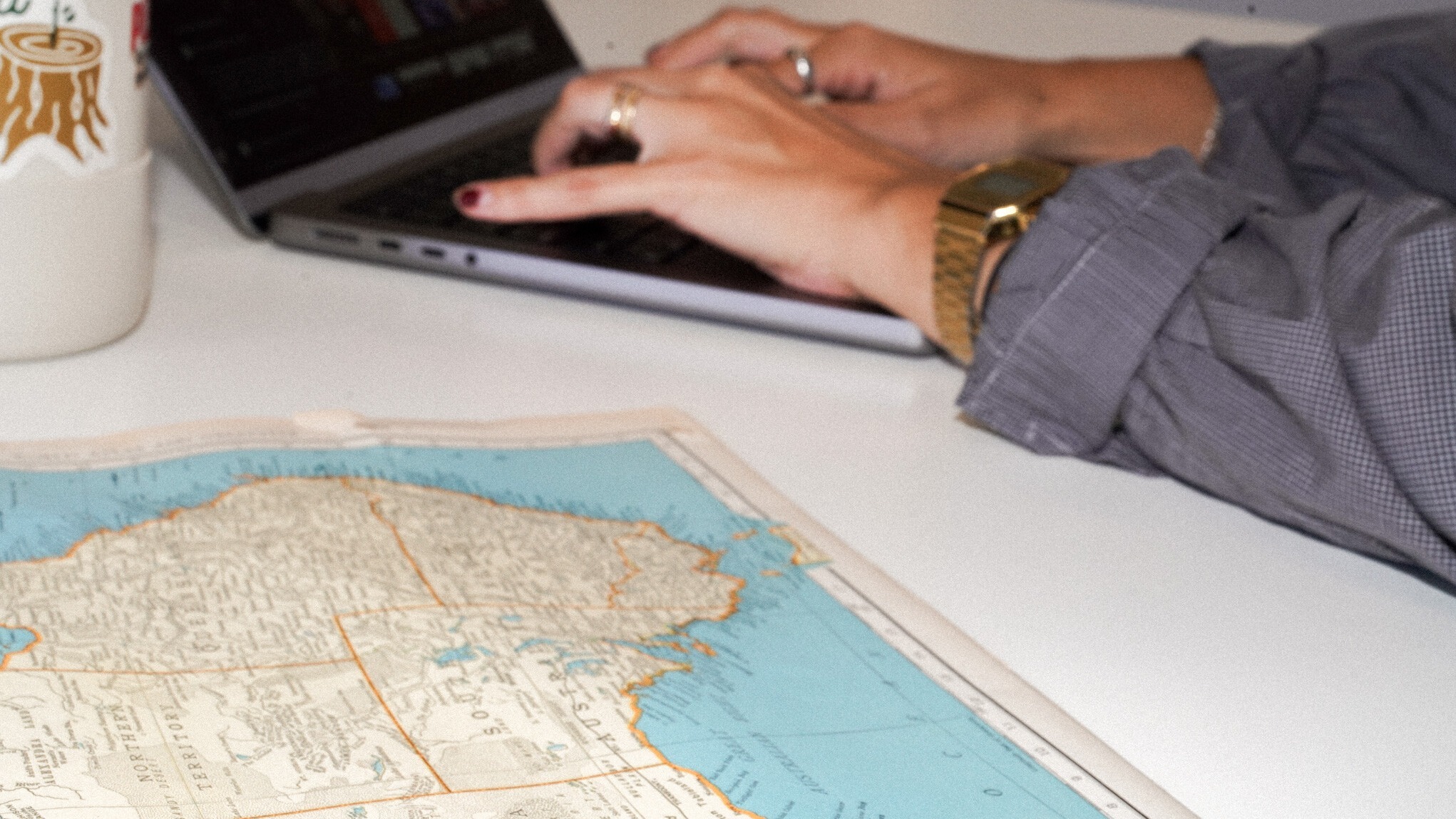 A person in a grey business shirt and gold watch types on a laptop computer at a white desk, with an Australian map spread out nearby.